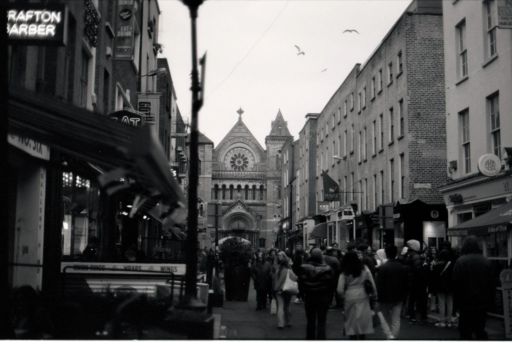 Black and white street scene showing pedestrians walking in front of storefronts with signs, with a historic church or cathedral with rose window and towers in the background and seagulls flying overhead.