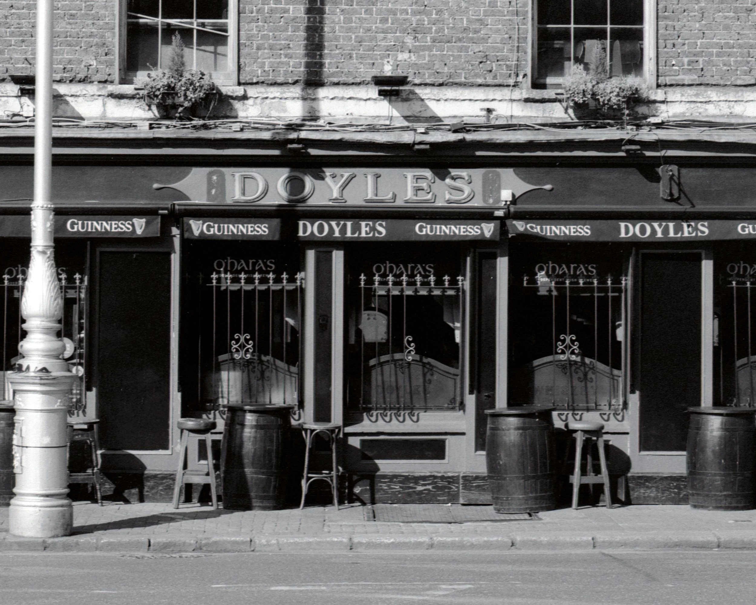 Facade of a pub called Doyles with signs for Guinness, outside seating with chairs, and a brick building with two windows and flower boxes.