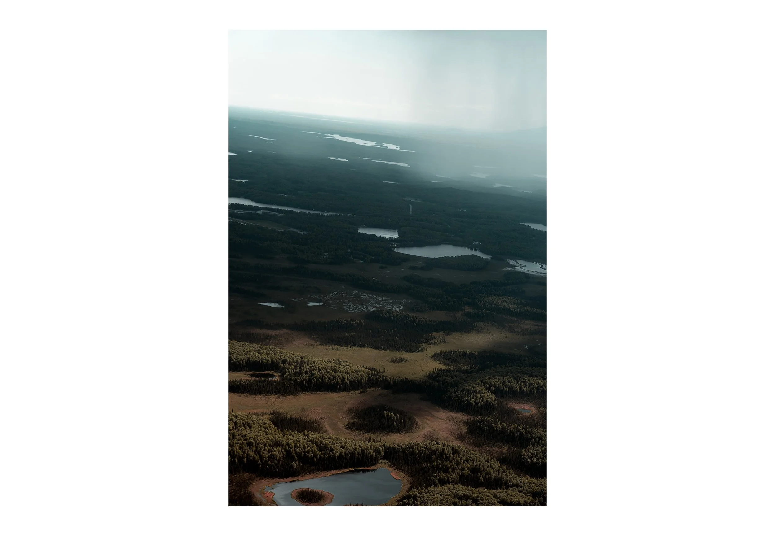 Aerial view of a landscape with multiple lakes surrounded by a dense forest, extending to the horizon under a hazy sky.