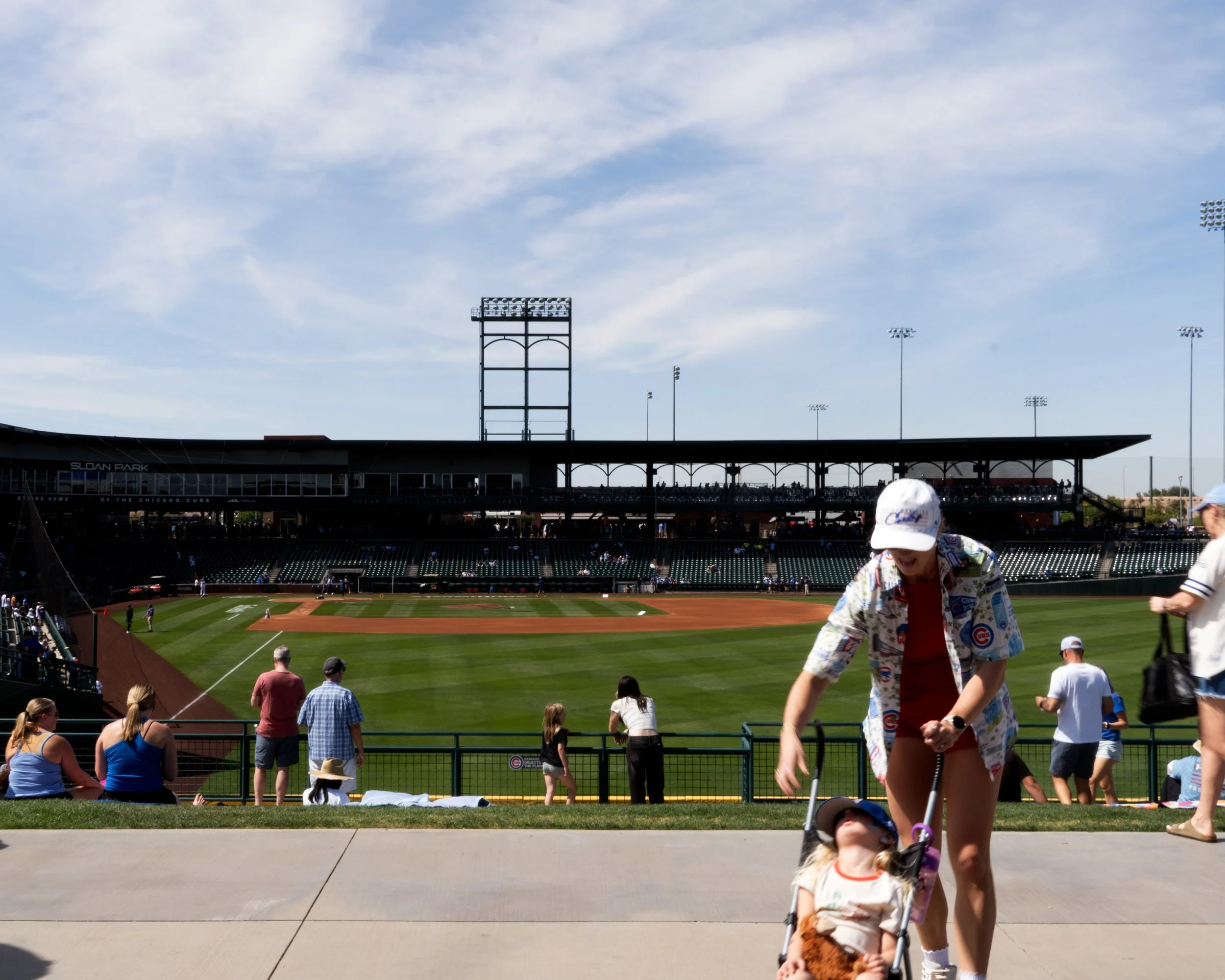 People walking and sitting in the stands of a baseball stadium on a sunny day, with some spectators and a baseball field in the background, and a woman in the foreground wearing a colorful shirt and a man wearing a white hat.