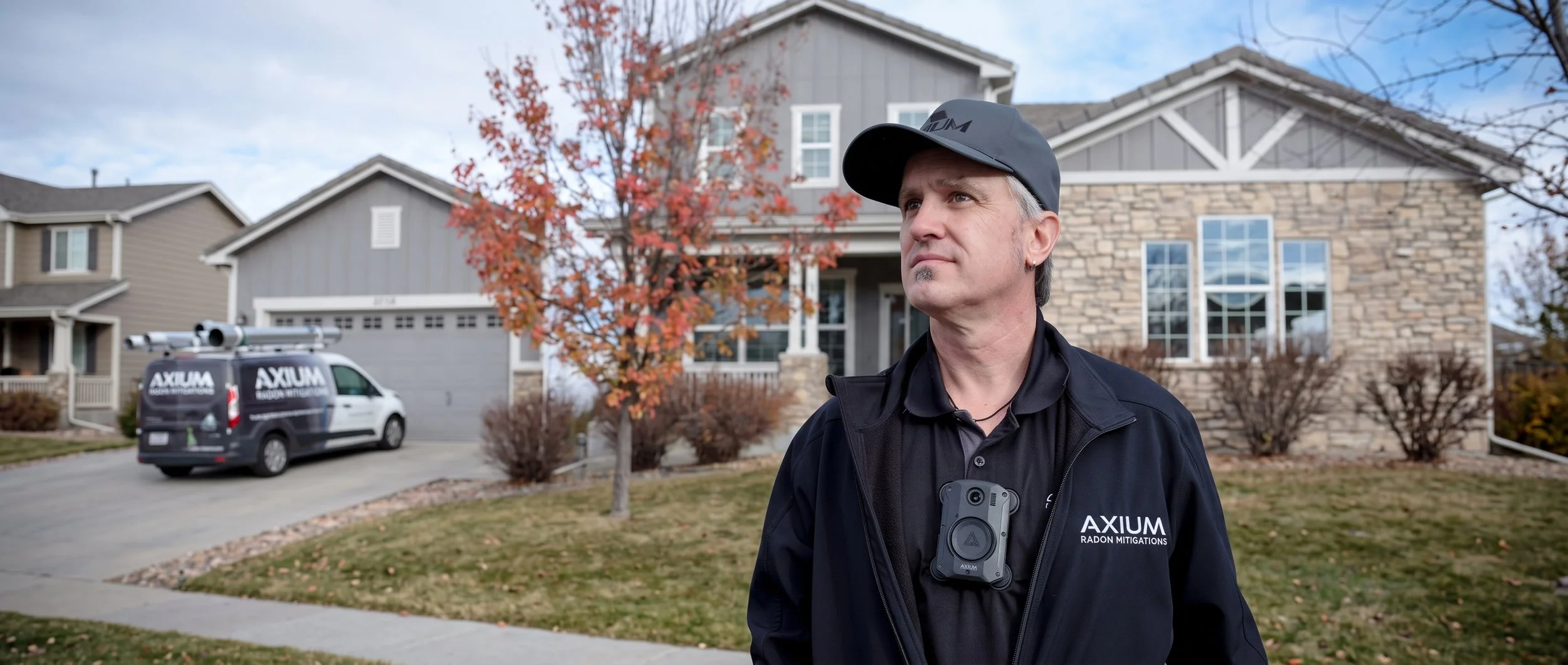 A man wearing a black jacket and cap with the brand 'AXIUM' standing outside in front of a two-story house. There is a company van labeled 'AXIUM Radon Mitigations' parked on the driveway, with a ladder mounted on top. The house has a gray exterior with white trim and a front porch, and autumn foliage is visible on a tree to the left.