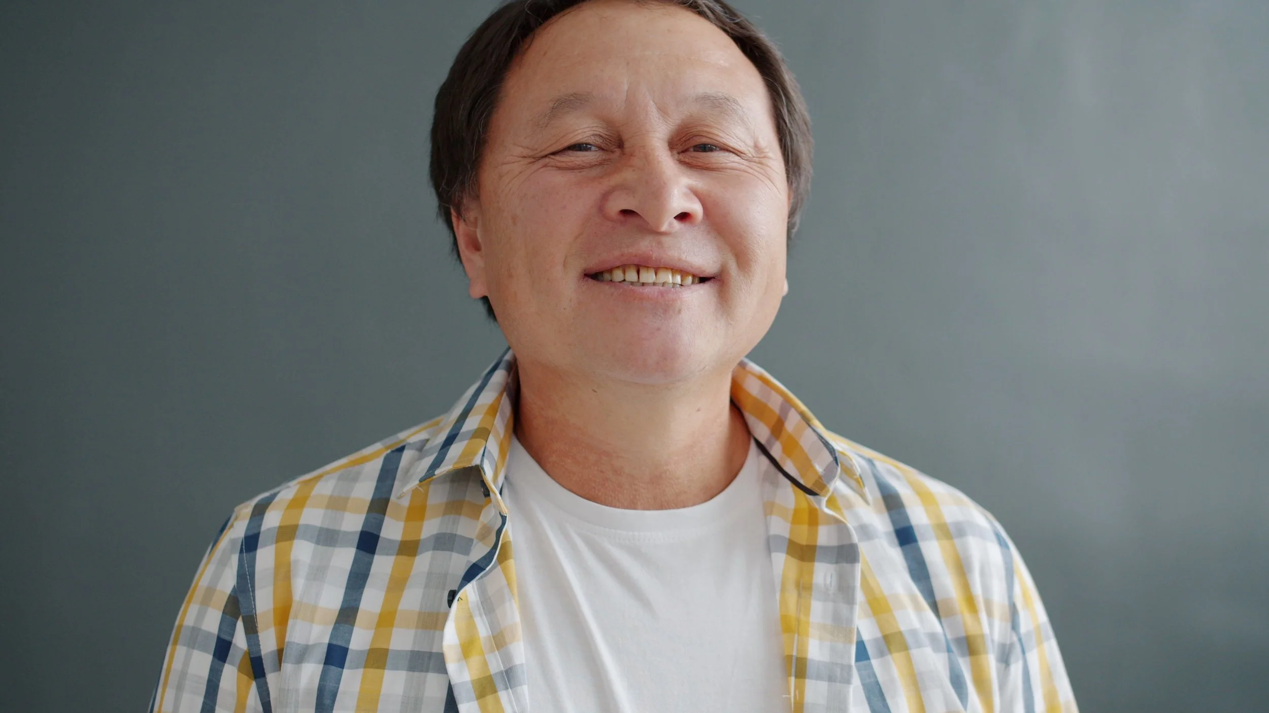 Close-up of a smiling young man with short brown hair wearing a yellow plaid shirt over a white t-shirt, standing against a plain gray background.
