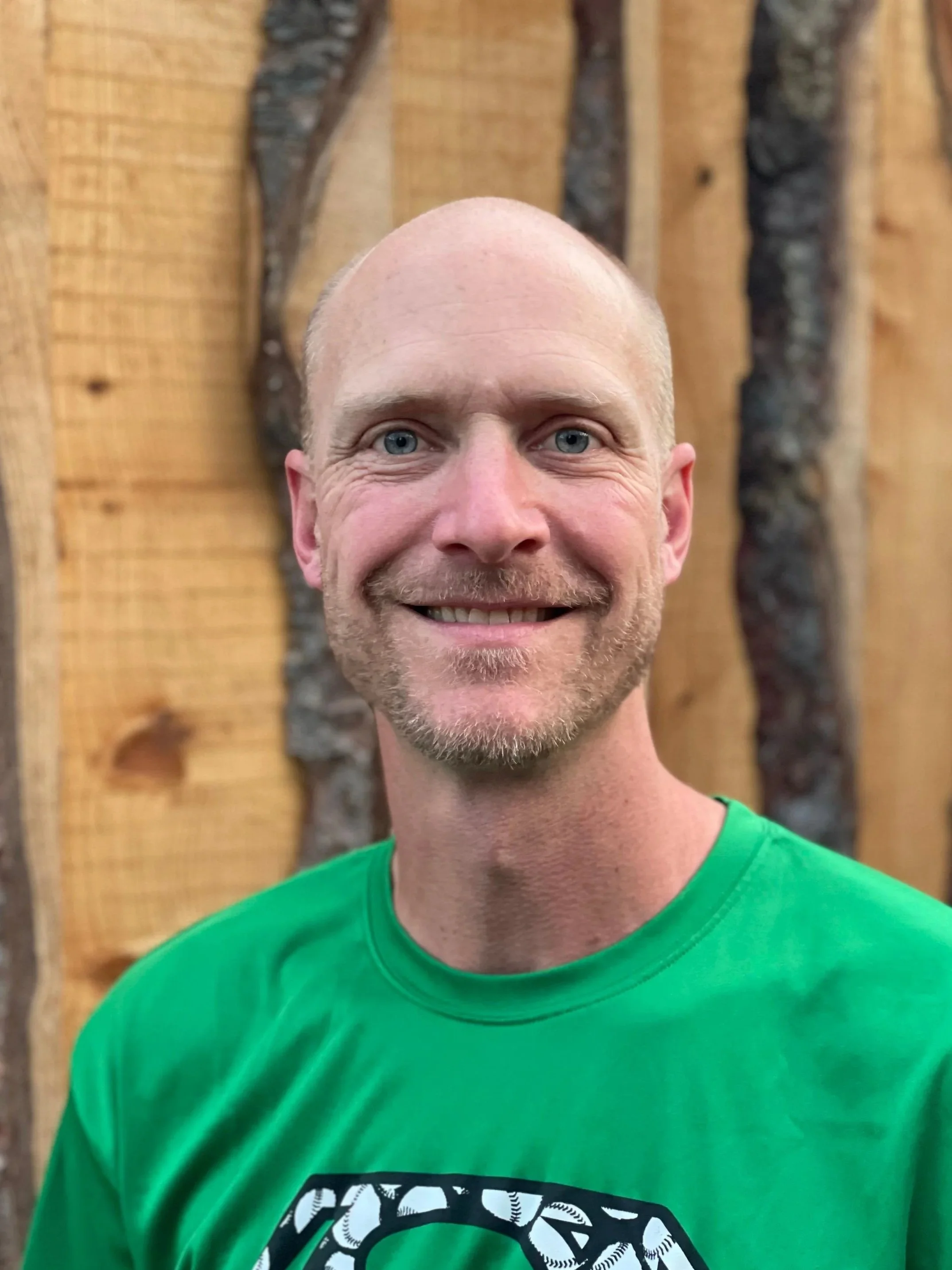 Close-up of a smiling man with a shaved head and blue eyes wearing a green shirt, standing in front of a wooden wall with bark accents.