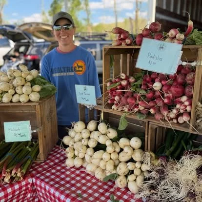 Smiling woman standing behind a table with baskets of radishes and onions at an outdoor farmers market.
