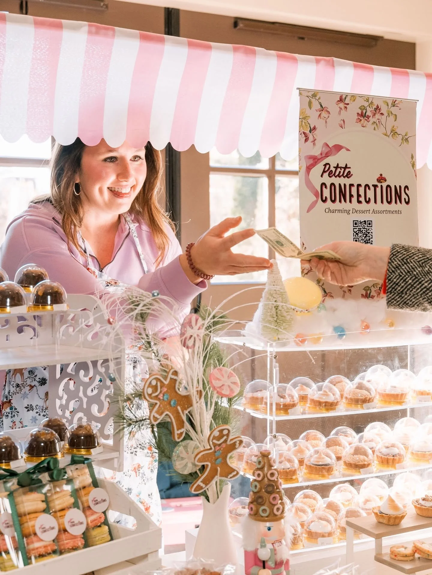 A woman smiling at a dessert stand displays a sign that reads 'Petite Confections' with colorful cupcakes and cookies, decorated for a festive holiday.
