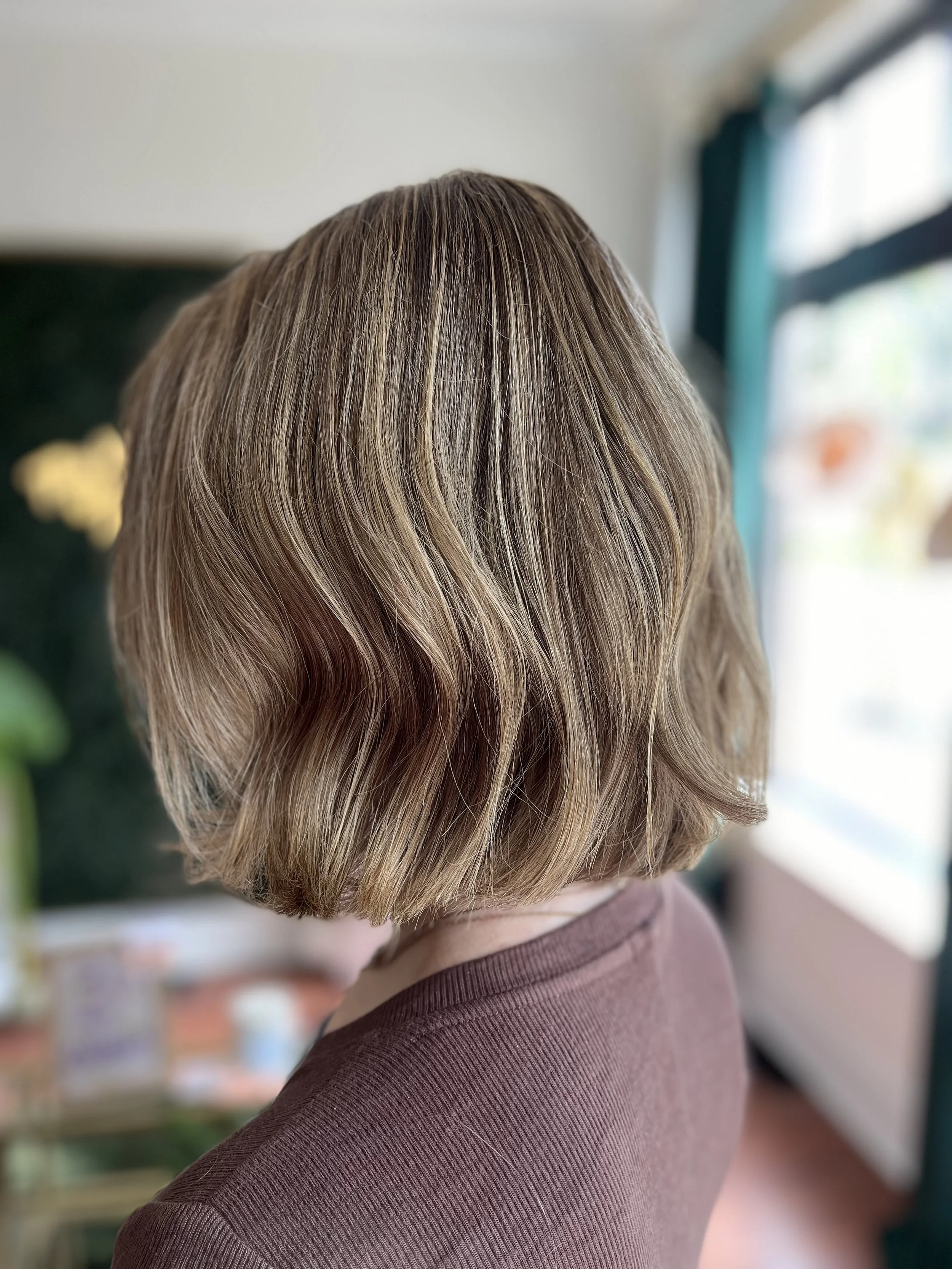 Back view of a woman with shoulder-length, light brown hair styled in loose waves, wearing a brown top, in a brightly lit room with blurred background.