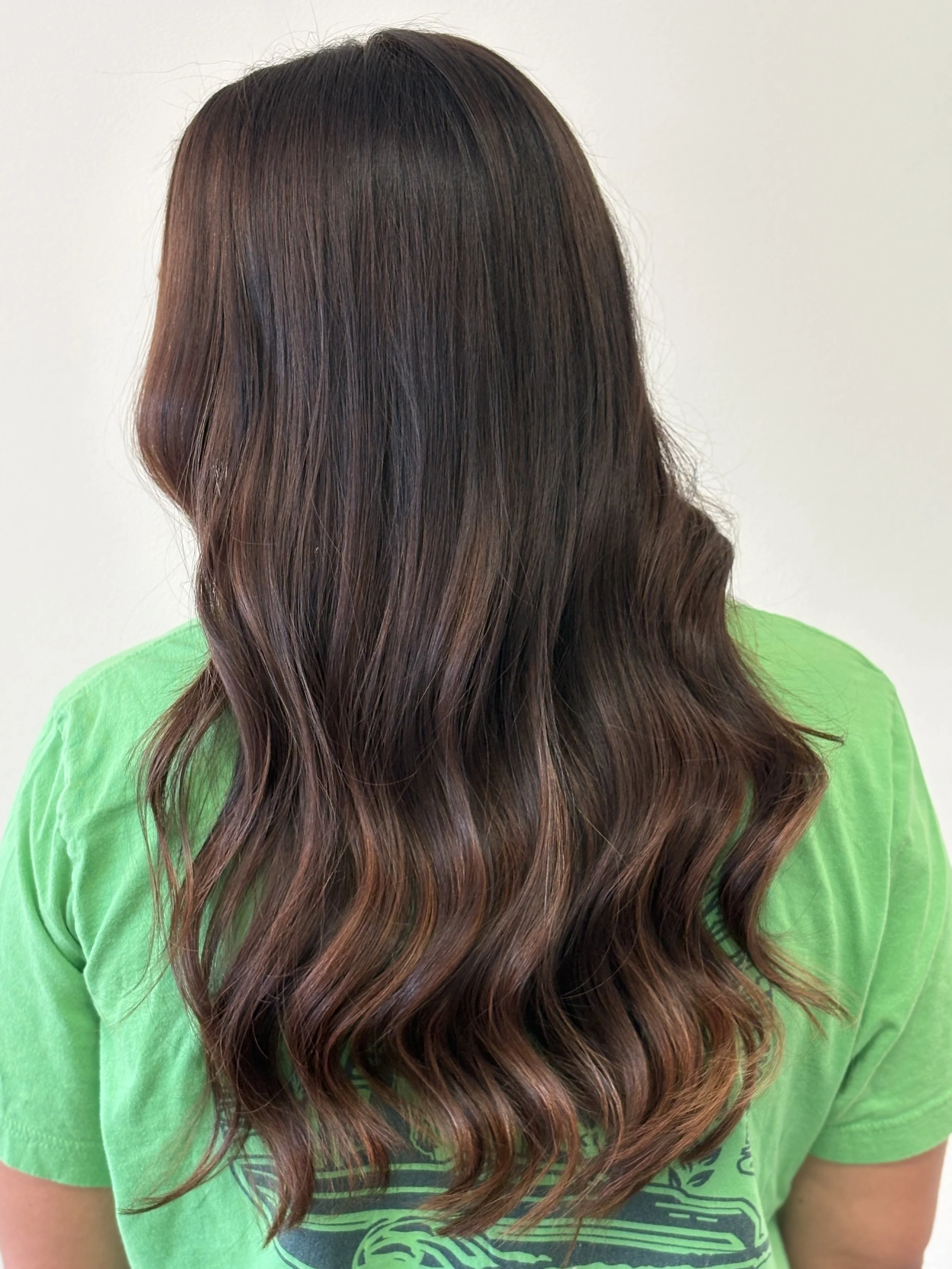 Person with long, wavy brown hair wearing a bright green T-shirt, facing away from the camera against a plain white background.
