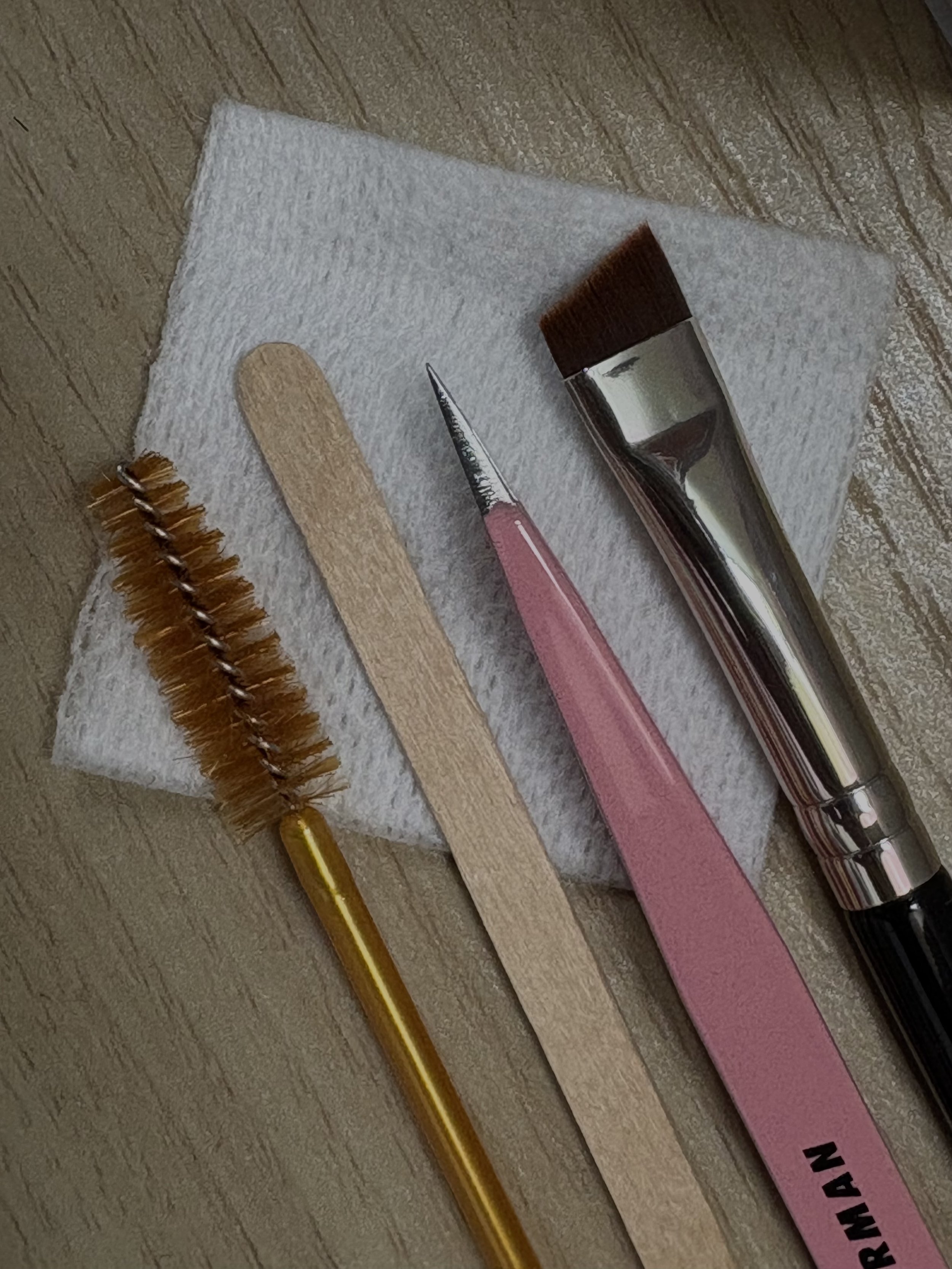 A close-up photo of three beauty tools for eyelash and eyebrow care on a white textured cloth: a gold eyelash brush, a wooden wax stick, pink tweezers, and a silver brow tint brush.