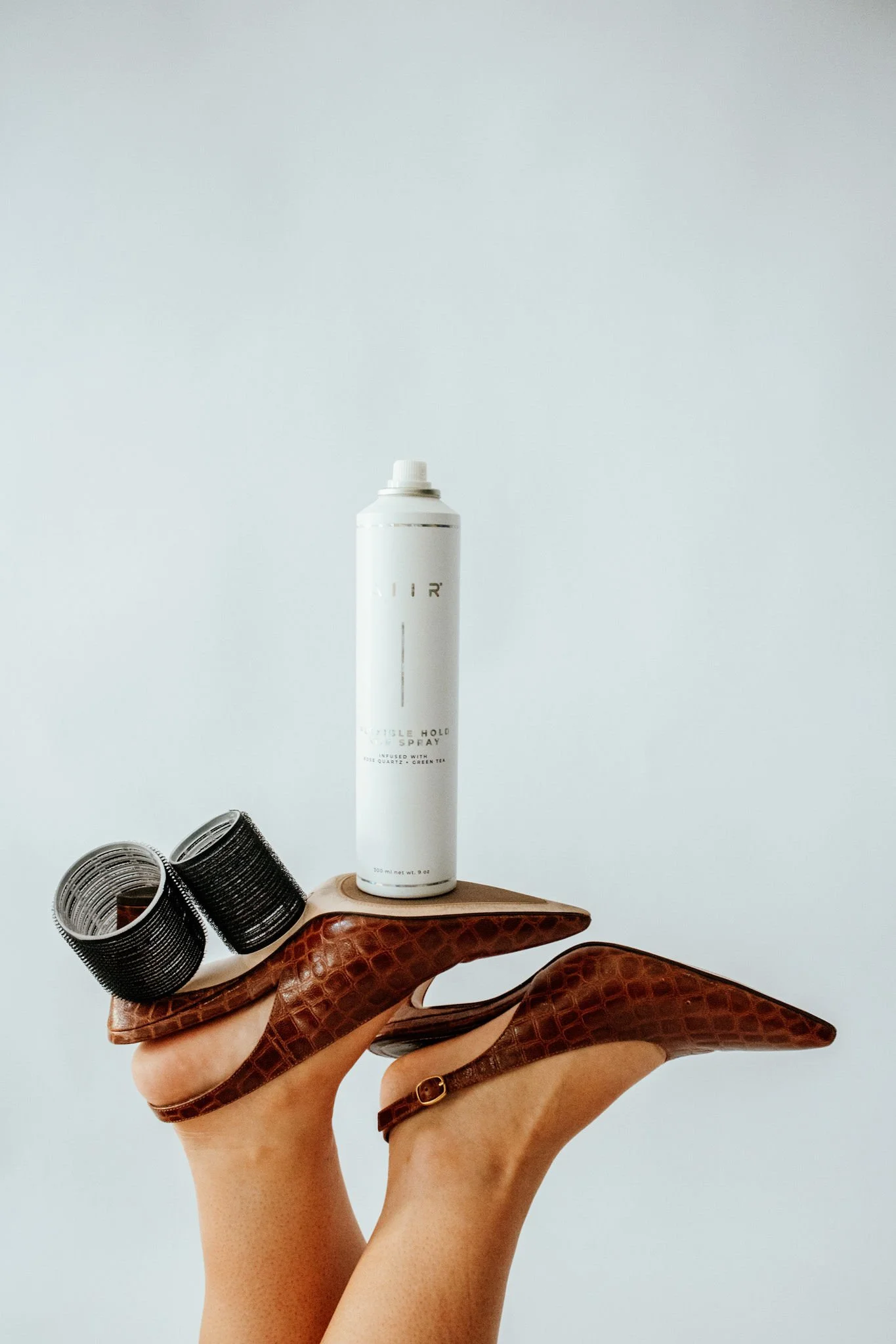 A hand holds a brown high-heeled shoe with black hair rollers and a white spray can balanced on the shoe's sole.