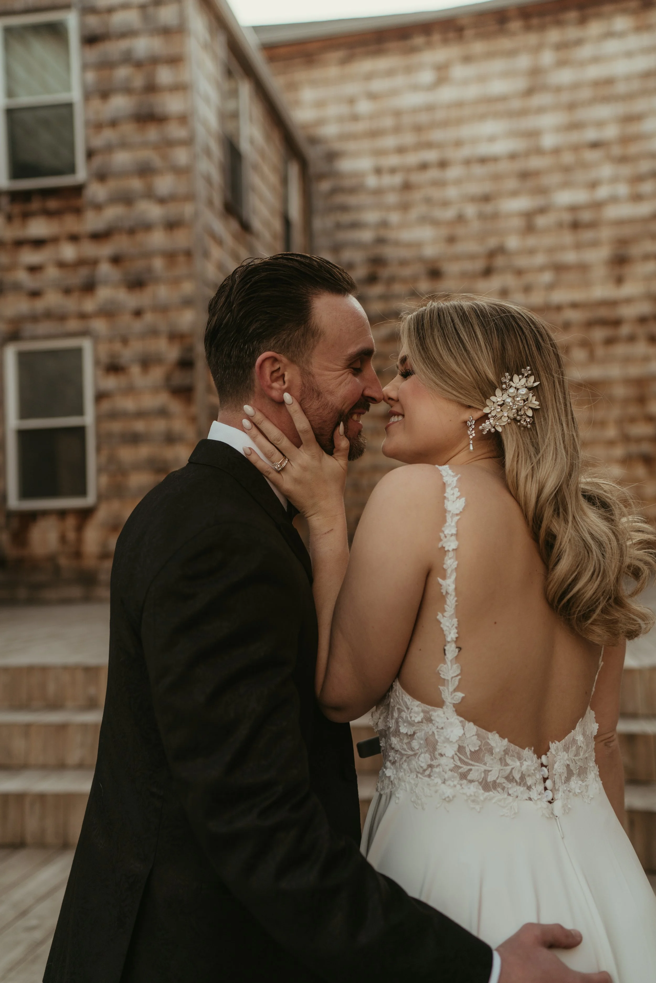 A bride and groom share a tender moment, standing close together, with their eyes closed and noses touching, in front of a rustic wooden building. The bride is wearing a lace wedding gown with floral details and a decorative hairpiece, while the groo