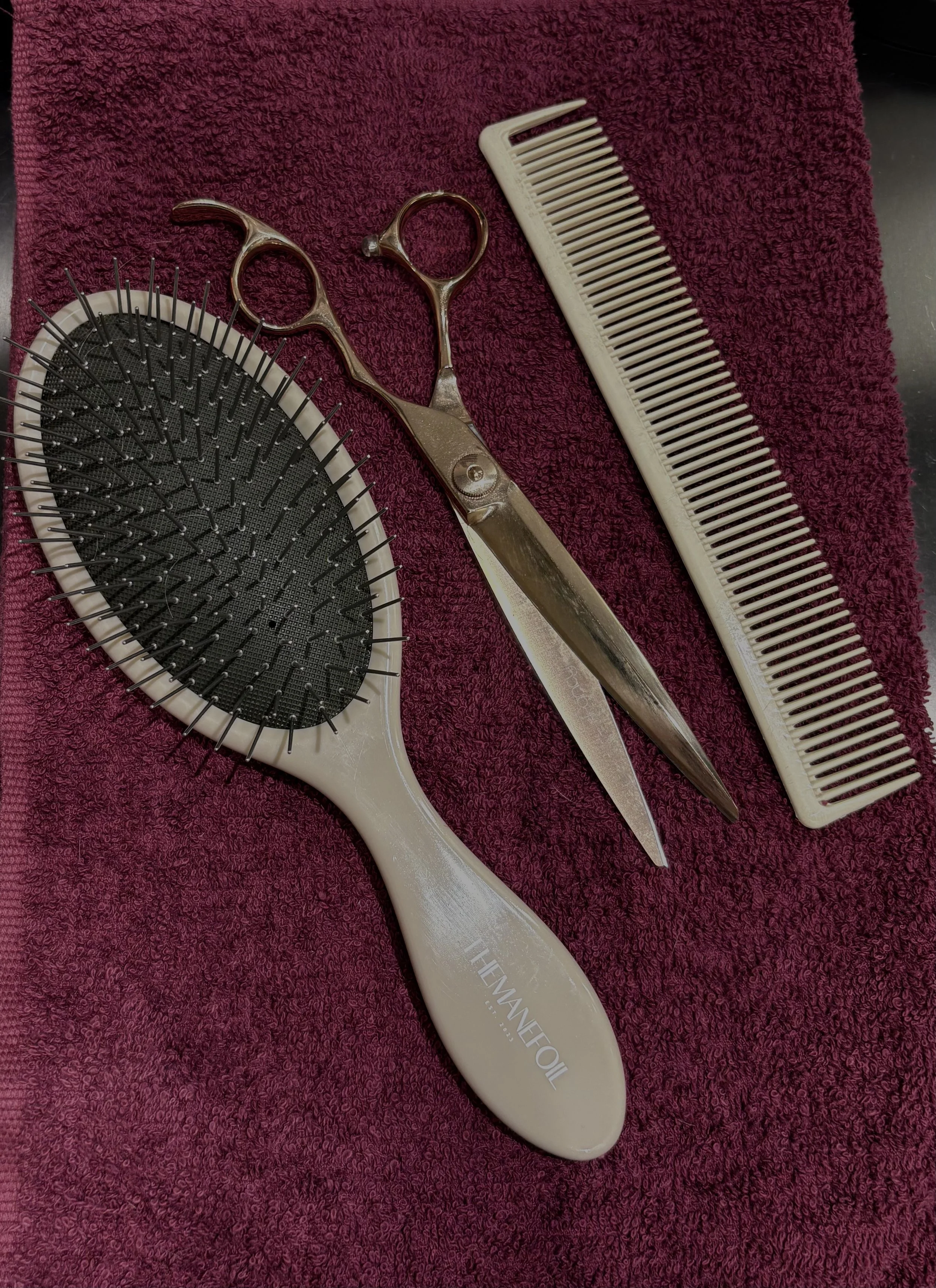 A white hairbrush with black bristles, a pair of metal scissors, and a white comb on a red towel.