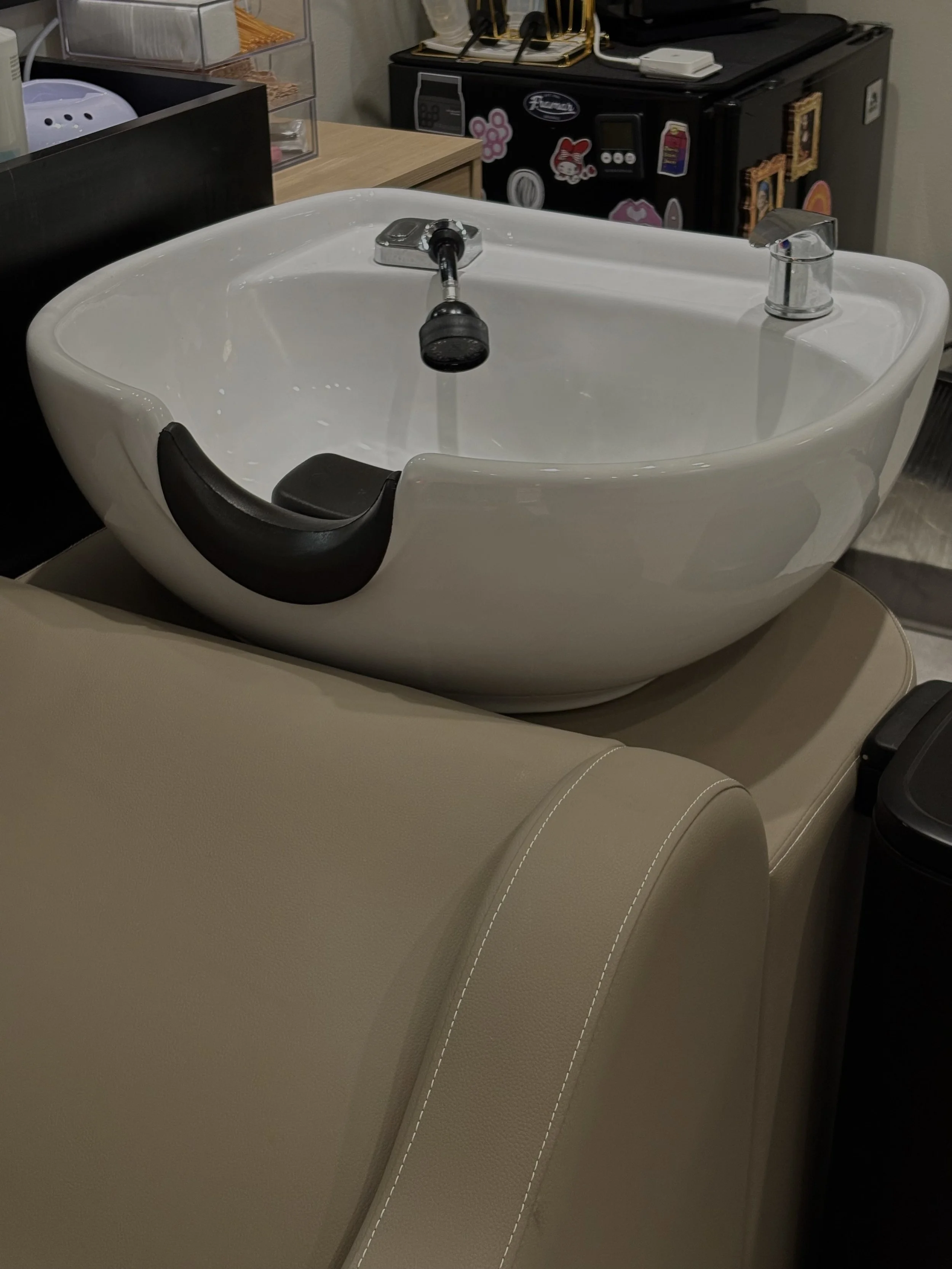 Close-up of a salon sink with a white porcelain basin, black cushion, and silver faucet and spray head, on a beige leather chair.