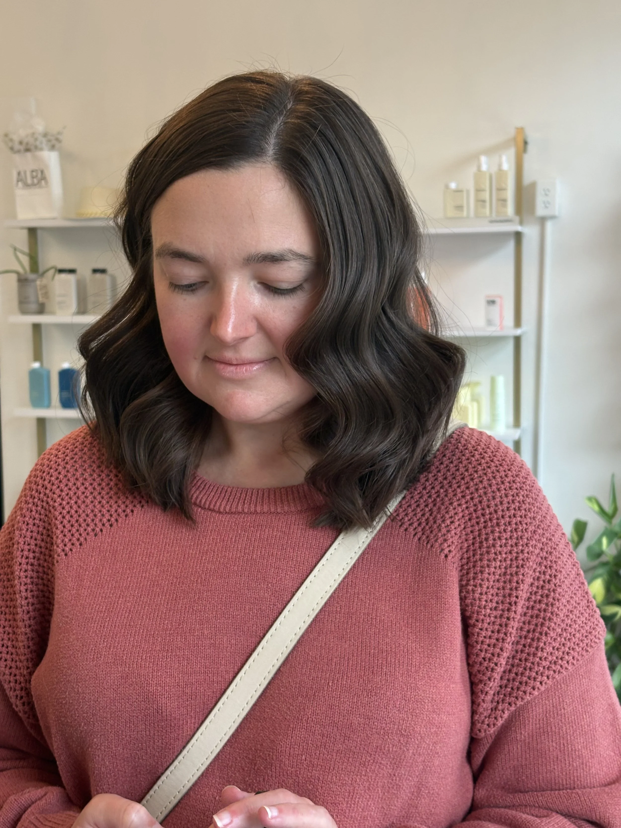 A woman with shoulder-length wavy dark brown hair and light skin wearing a red knit sweater and a white strap on her shoulder, standing in front of a white bookshelf with skincare and hair products and a small plant, in a bright room.