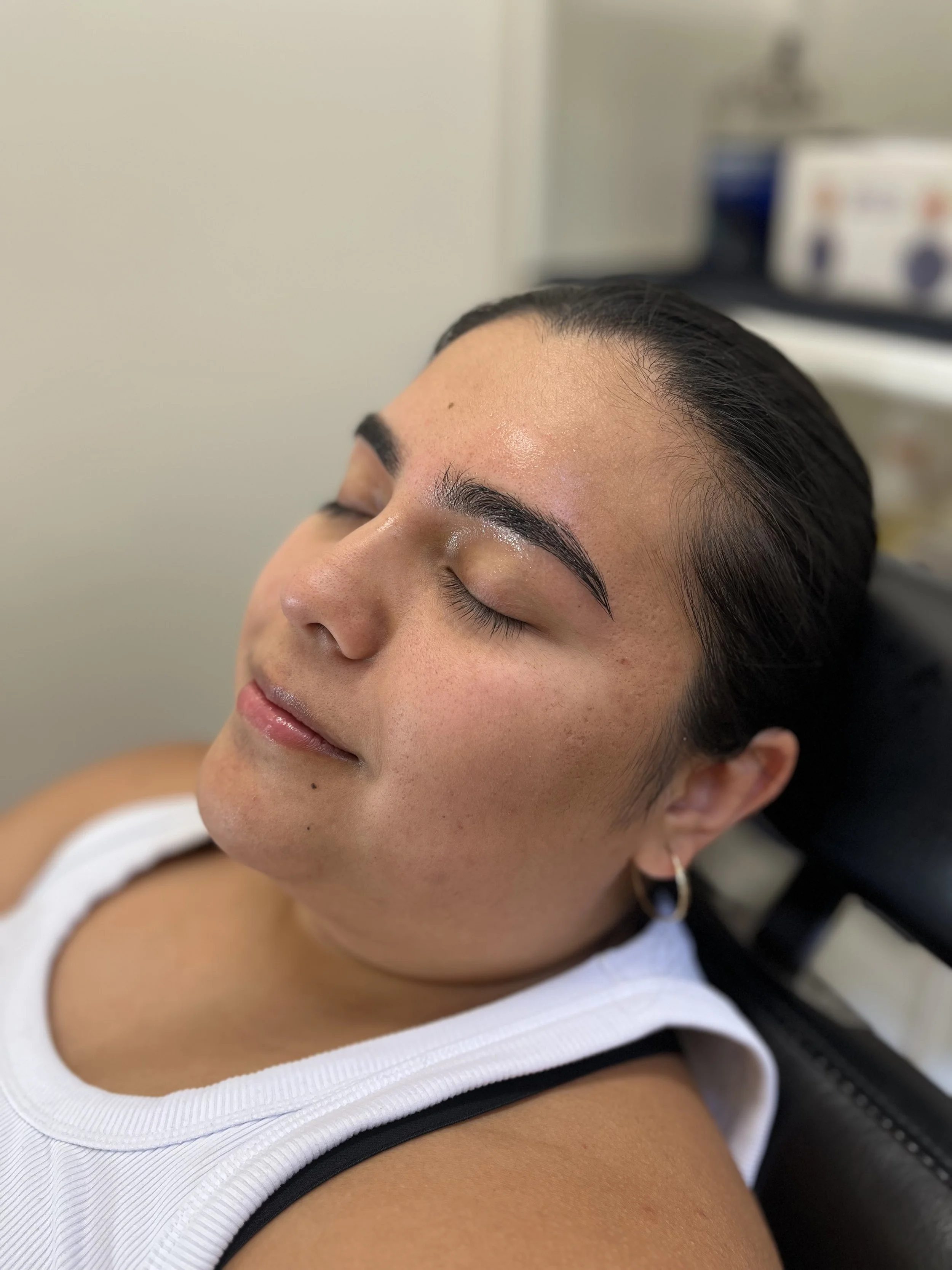 A woman with dark hair and earrings lying with her eyes closed, with a slight smile, in a clinical setting.