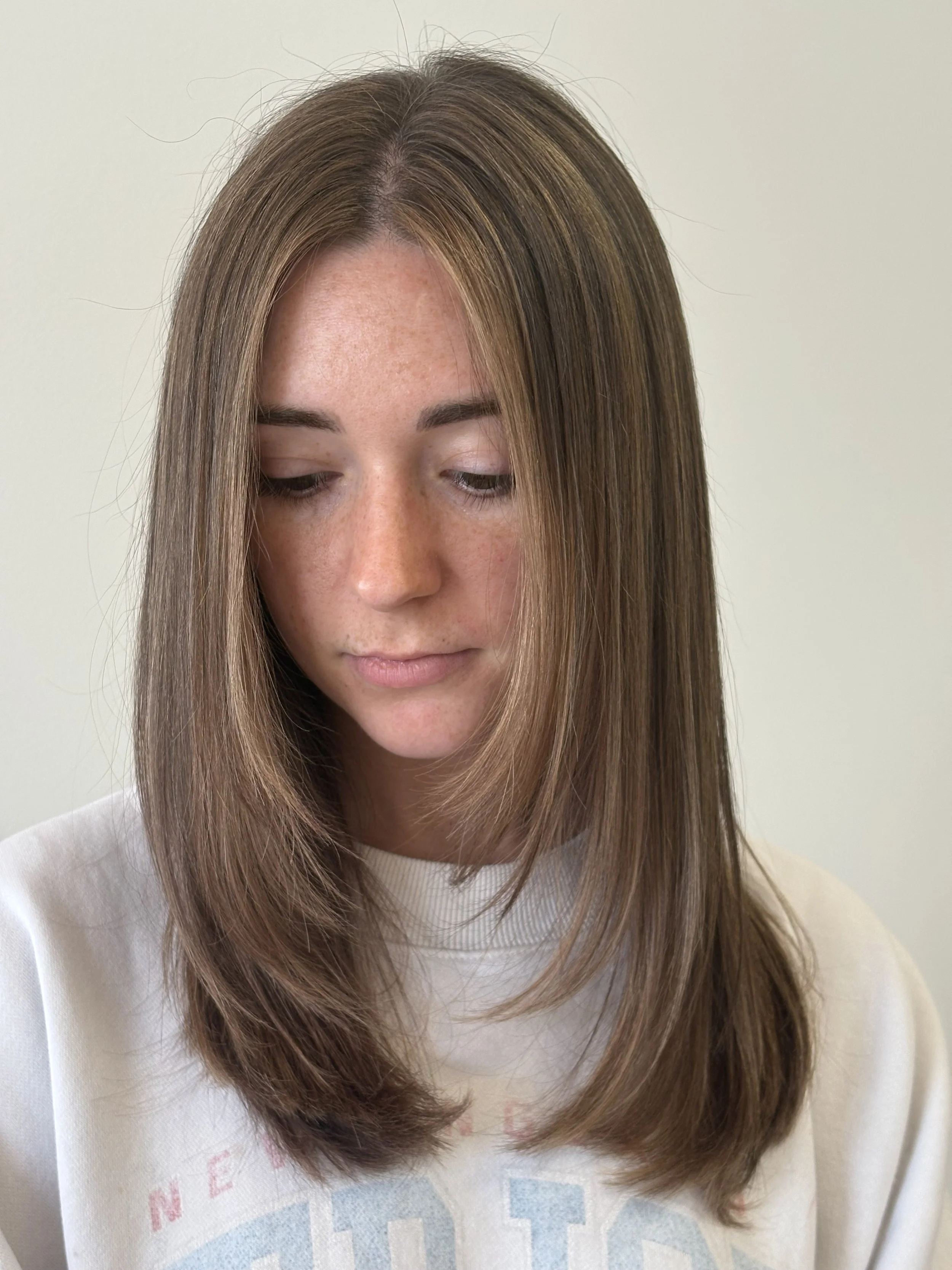Close-up of a woman with long, straight, light brown hair with highlights, wearing a white shirt with a colorful graphic and the word 'NEED' visible. She is looking down with her eyes closed, and the background is a plain, light-colored wall.