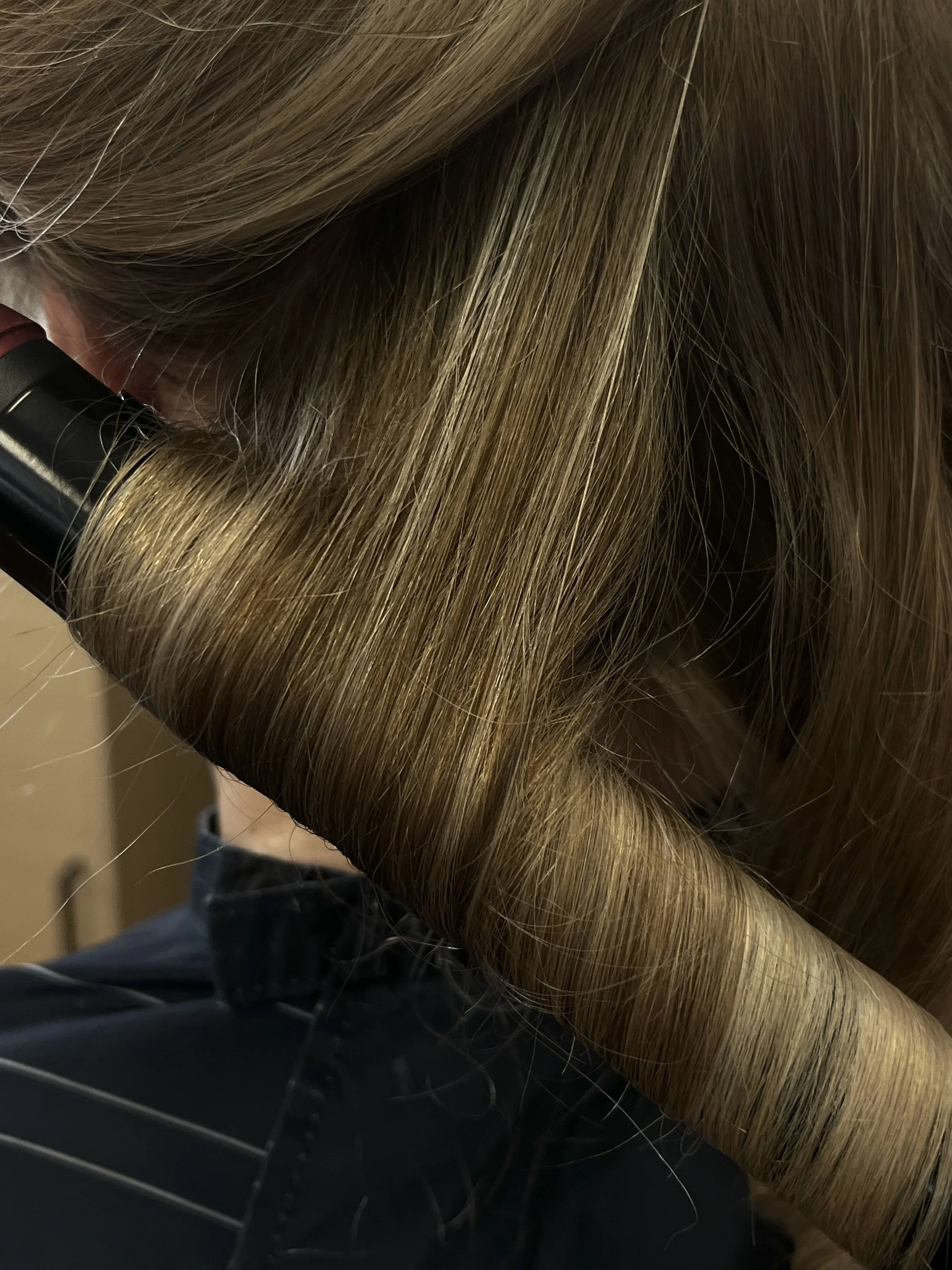 Close-up of a person with long, straight, light brown hair using a curling iron on their hair.