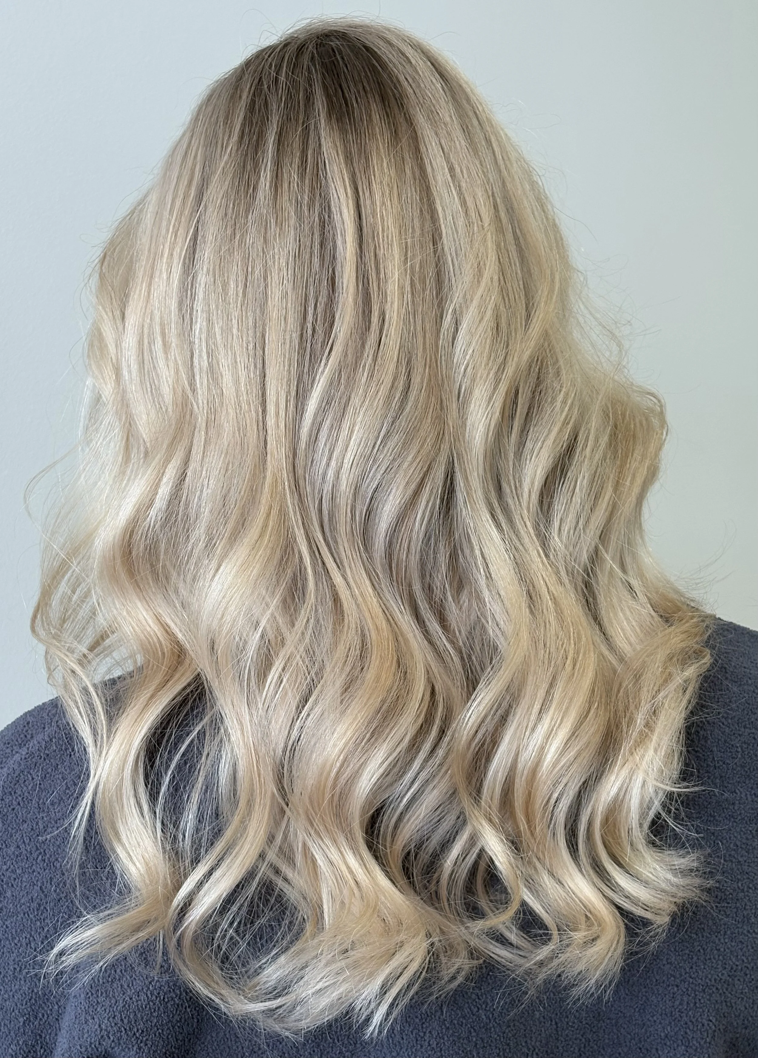 Back view of long wavy blonde hair with subtle lowlights, worn over a dark sweater, against a light gray background.