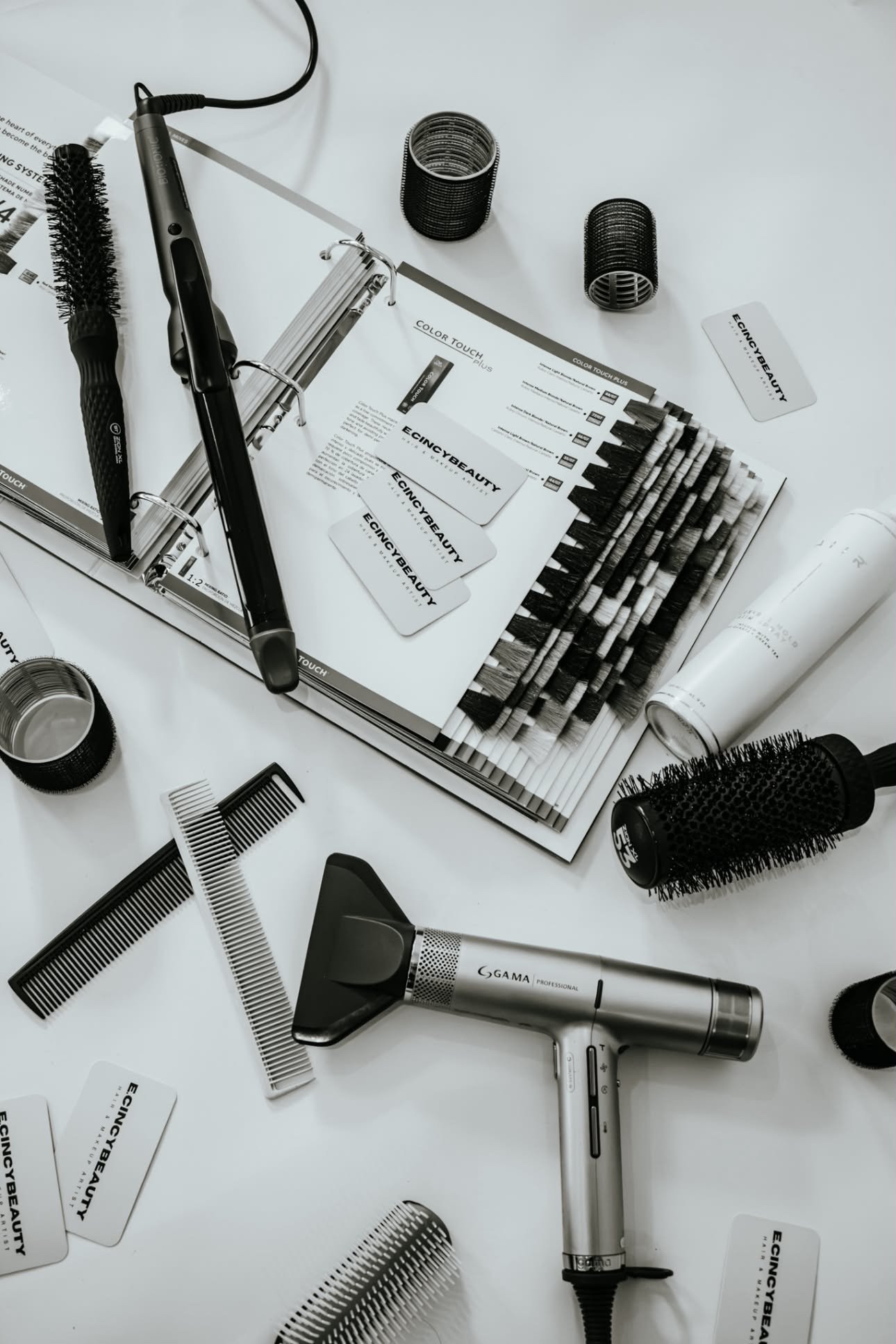 Various hair styling tools and accessories on a white table, including a curling iron, hairbrushes, combs, hair rollers, and promotional cards for Ecincy Beauty.