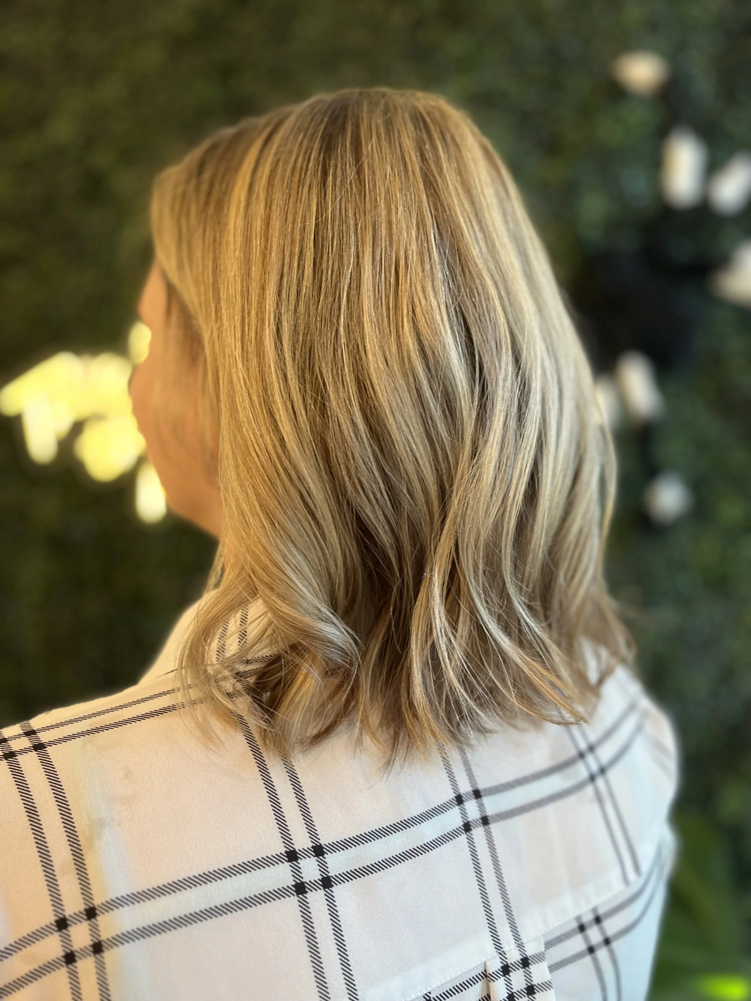 Woman with long, wavy blonde hair wearing a plaid blazer with black lines on a white background, standing outdoors with blurred greenery and white flowers in the background.