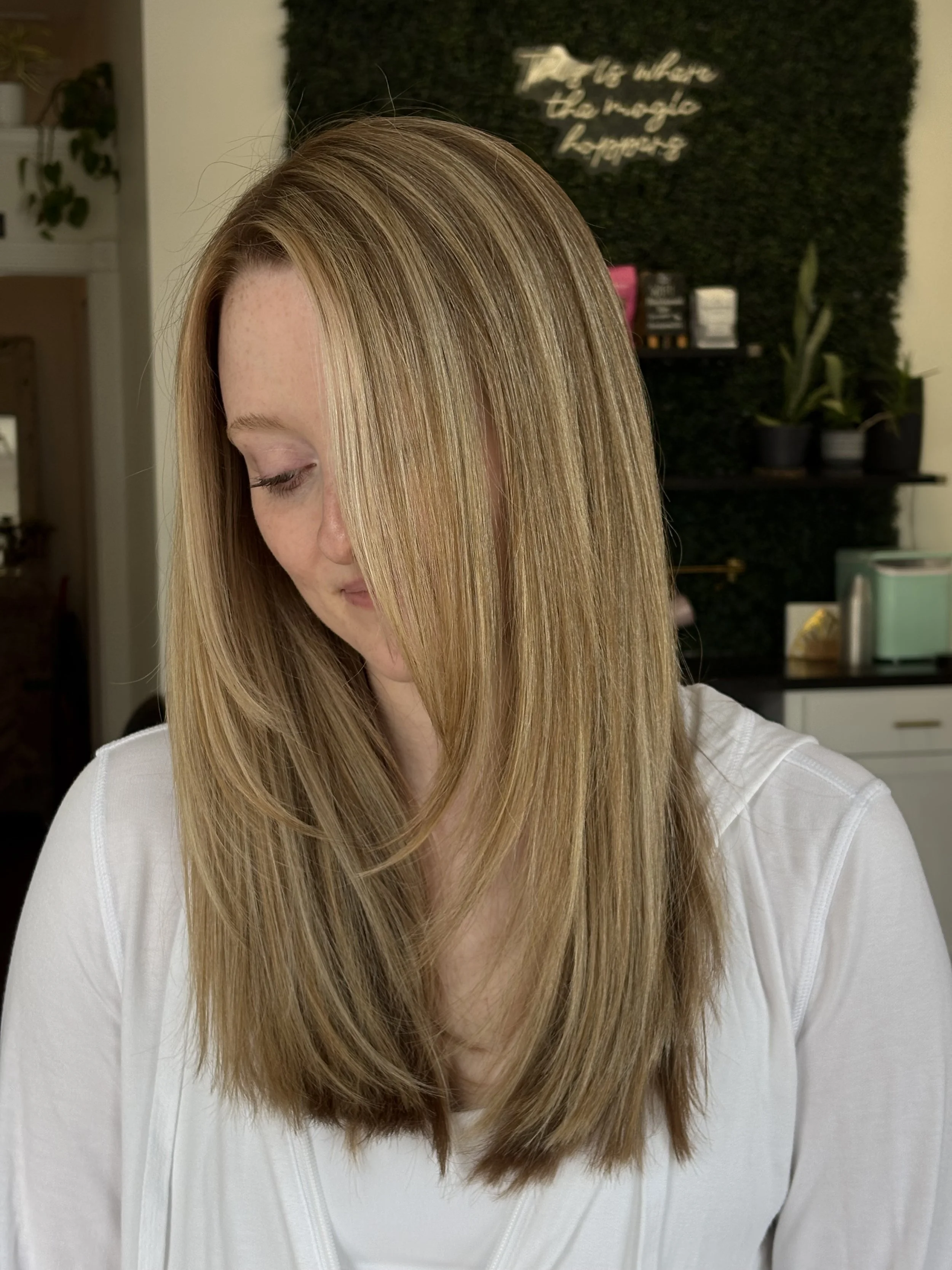 Close-up of a woman with long, straight blonde hair, wearing a white top, with a blurred indoor background including a black wall with plants and shelves.