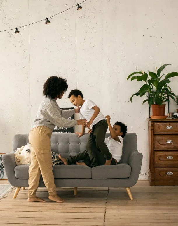 A woman and two young boys playing and laughing on a gray sofa in a living room with a white wall, string lights overhead, a potted plant on a wooden side table, and a wooden floor.