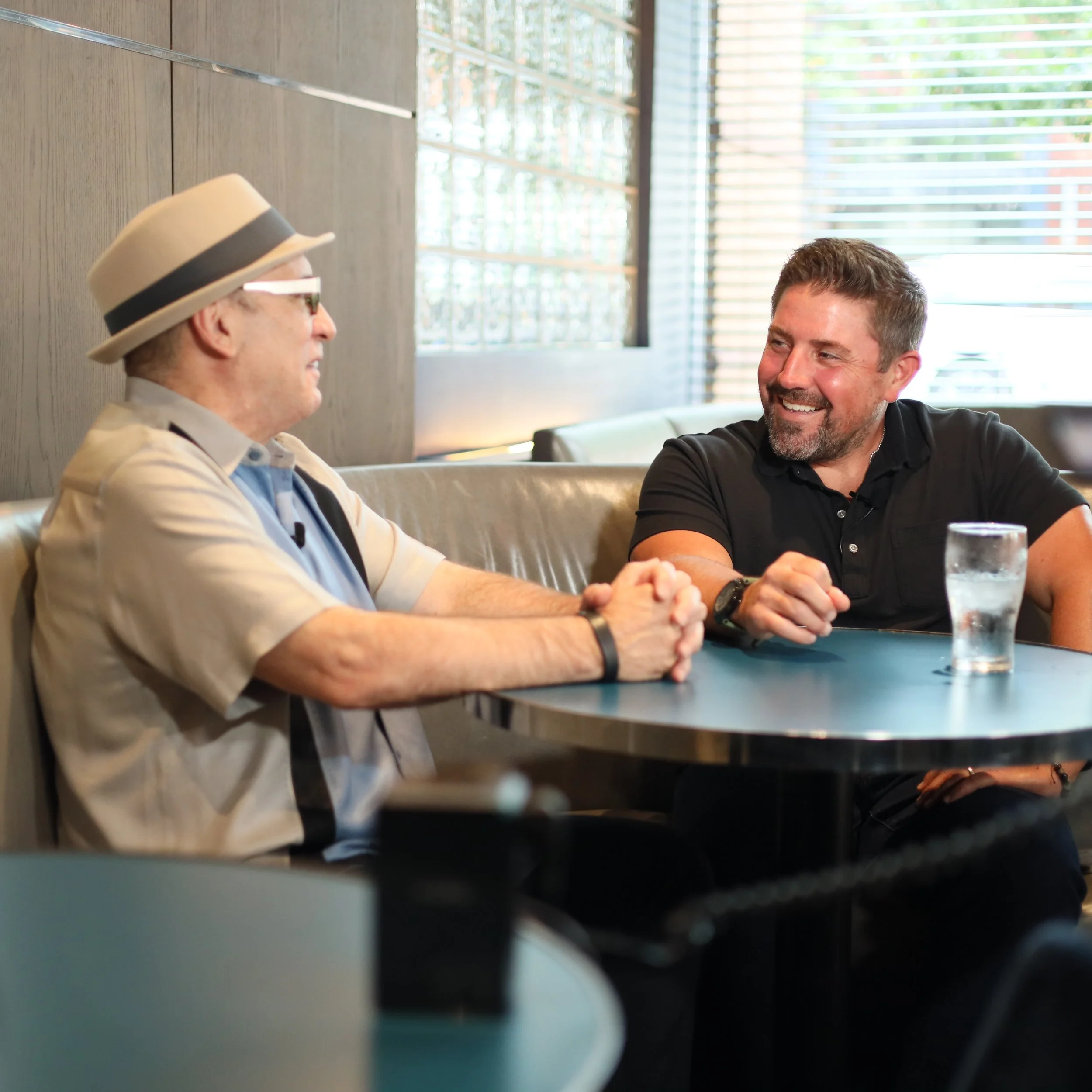 Two men sitting at a table, smiling and engaging in conversation, with natural light coming through the window blinds behind them.