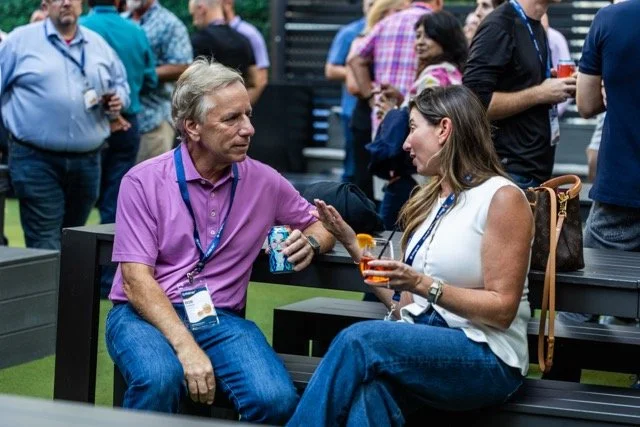 Two people, a man and a woman, are talking and smiling at a social gathering outdoors. The man is sitting on a bench, wearing a purple shirt and jeans. The woman is sitting on a similar bench, wearing a white top and jeans, holding a drink, and gesturing as they converse.