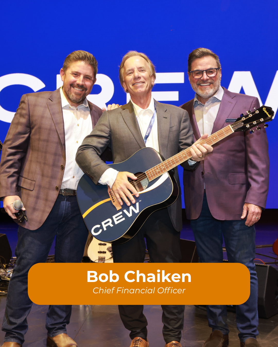 Three men on stage at a corporate event, with one holding a guitar labeled 'CREW' and another holding a microphone. The background features partial text, and a caption reads 'Bob Chaiken, Chief Financial Officer.'