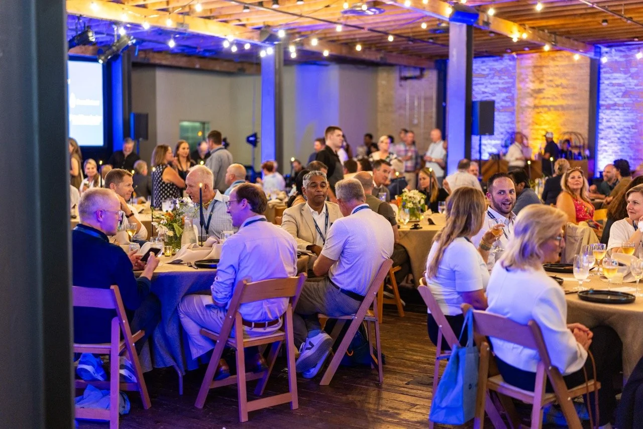 People seated at round tables with white tablecloths and floral centerpieces in a decorated event space with string lights and brick walls, attending a social gathering or dinner event.