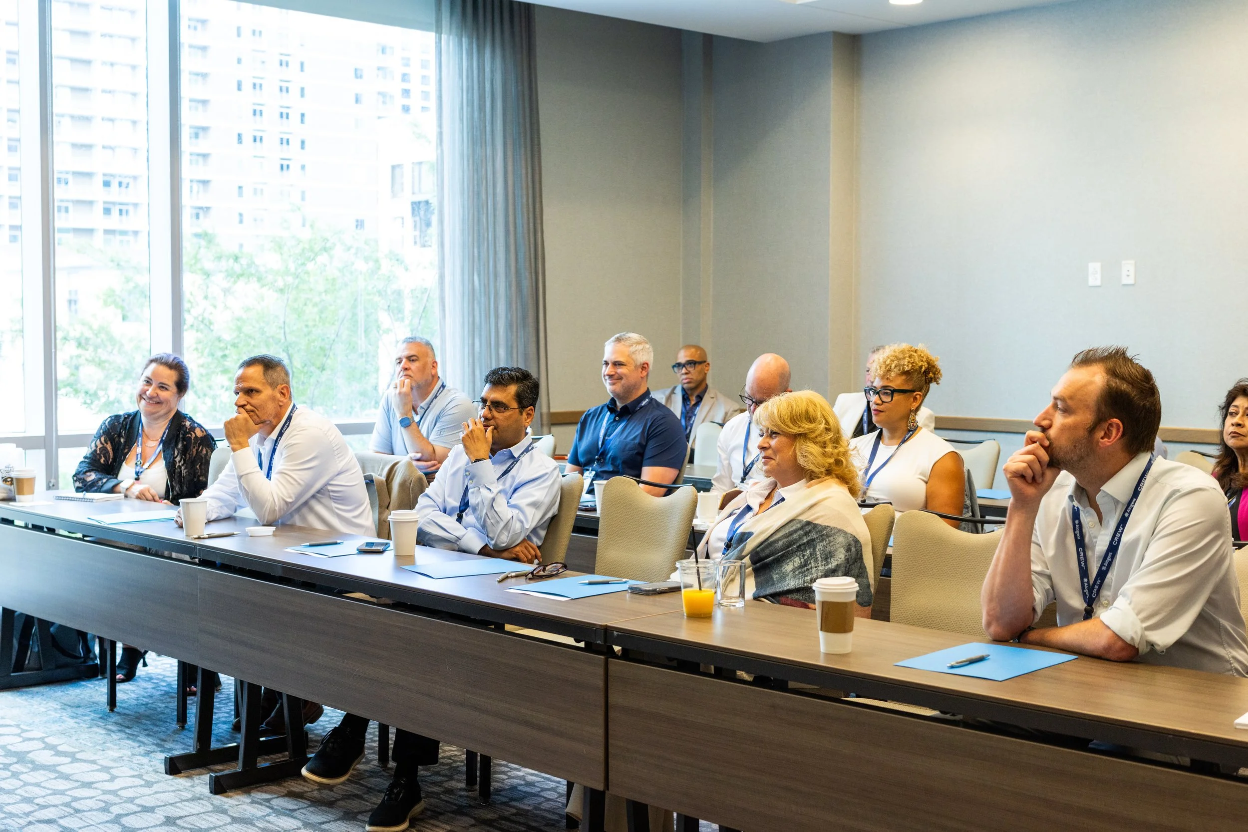 People attending a conference or seminar in a bright meeting room with large windows. They are seated at long tables, some with notebooks, pens, and beverages in front of them, listening attentively.