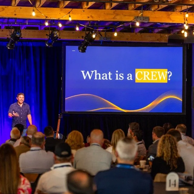 A presenter on stage at a conference, standing in front of a large screen that says 'What is a CREW?' in a room with an audience. The room has wooden ceilings and string lights.