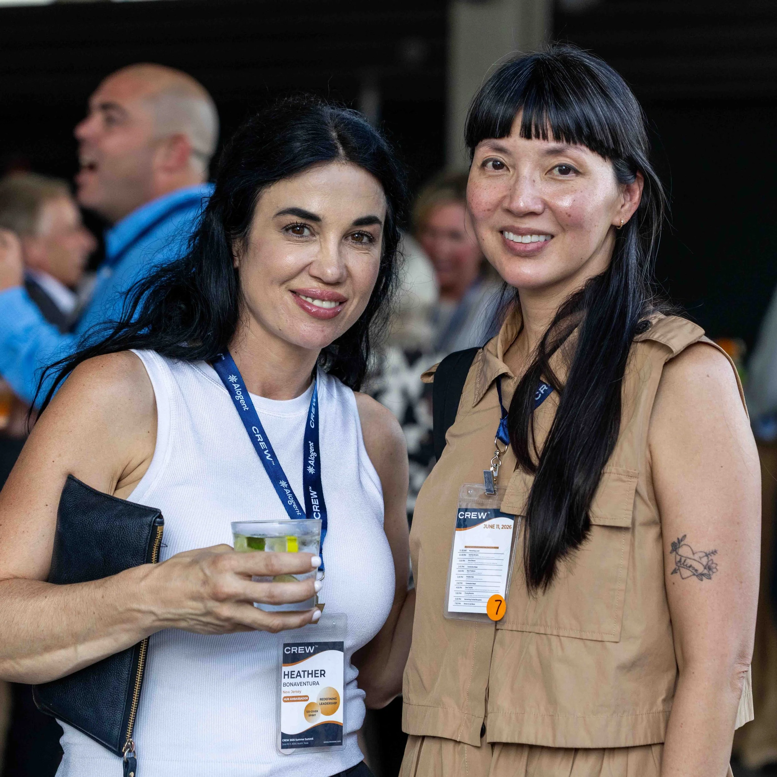 Two women smiling at the camera at a conference, wearing name badges and conference lanyards, with other people in the background.