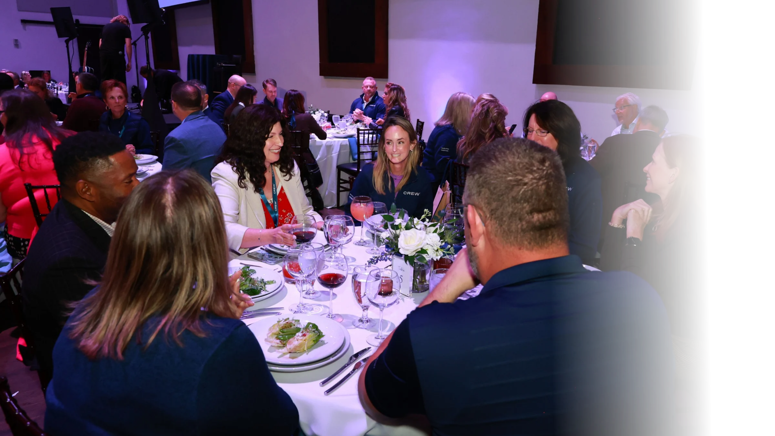 People sitting at round tables with white tablecloths, wine glasses, and floral centerpieces, engaged in conversation at a formal event or dinner.