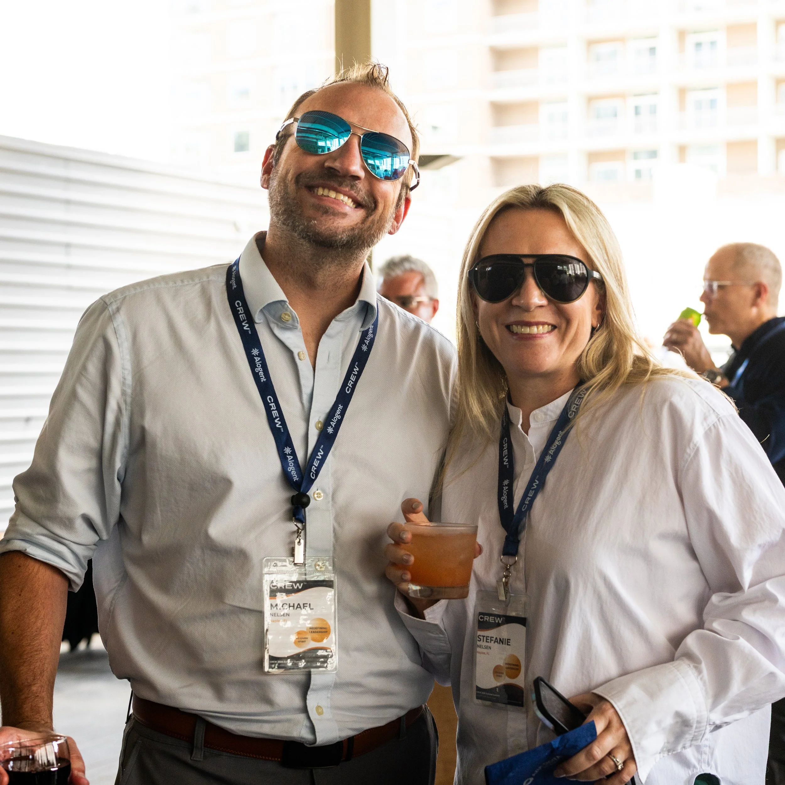 Two people smiling for a photo at a professional event, wearing name badges on lanyards, with one holding a glass of red wine and the other holding a cup of coffee.