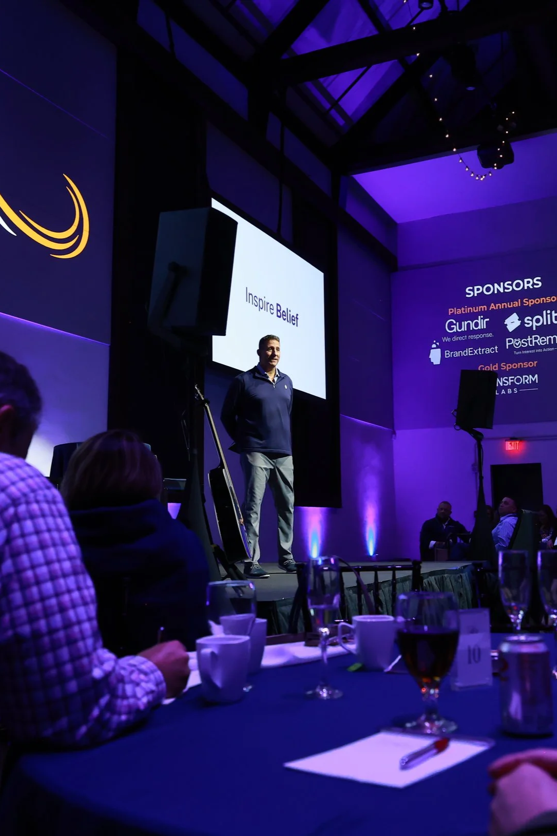 Man giving a presentation on stage at a conference with a large screen behind him displaying the words 'Inspire Belief'. Audience members seated at tables are visible, with glasses and cups on the tables, in a dimly lit room with purple lighting.