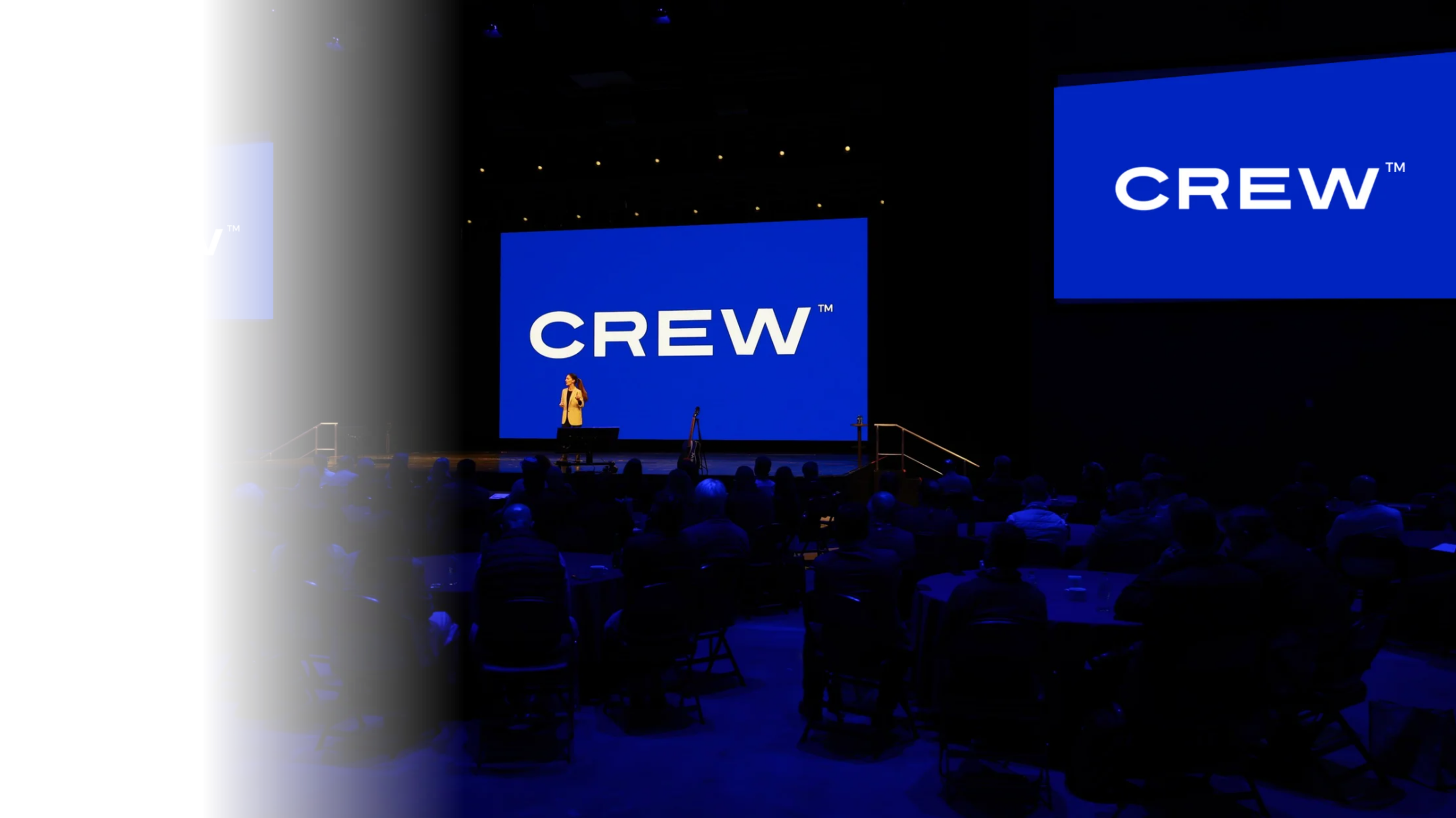 A person standing on stage in front of large blue screens displaying the word 'CREW' during a presentation or conference, with an audience seated in front.