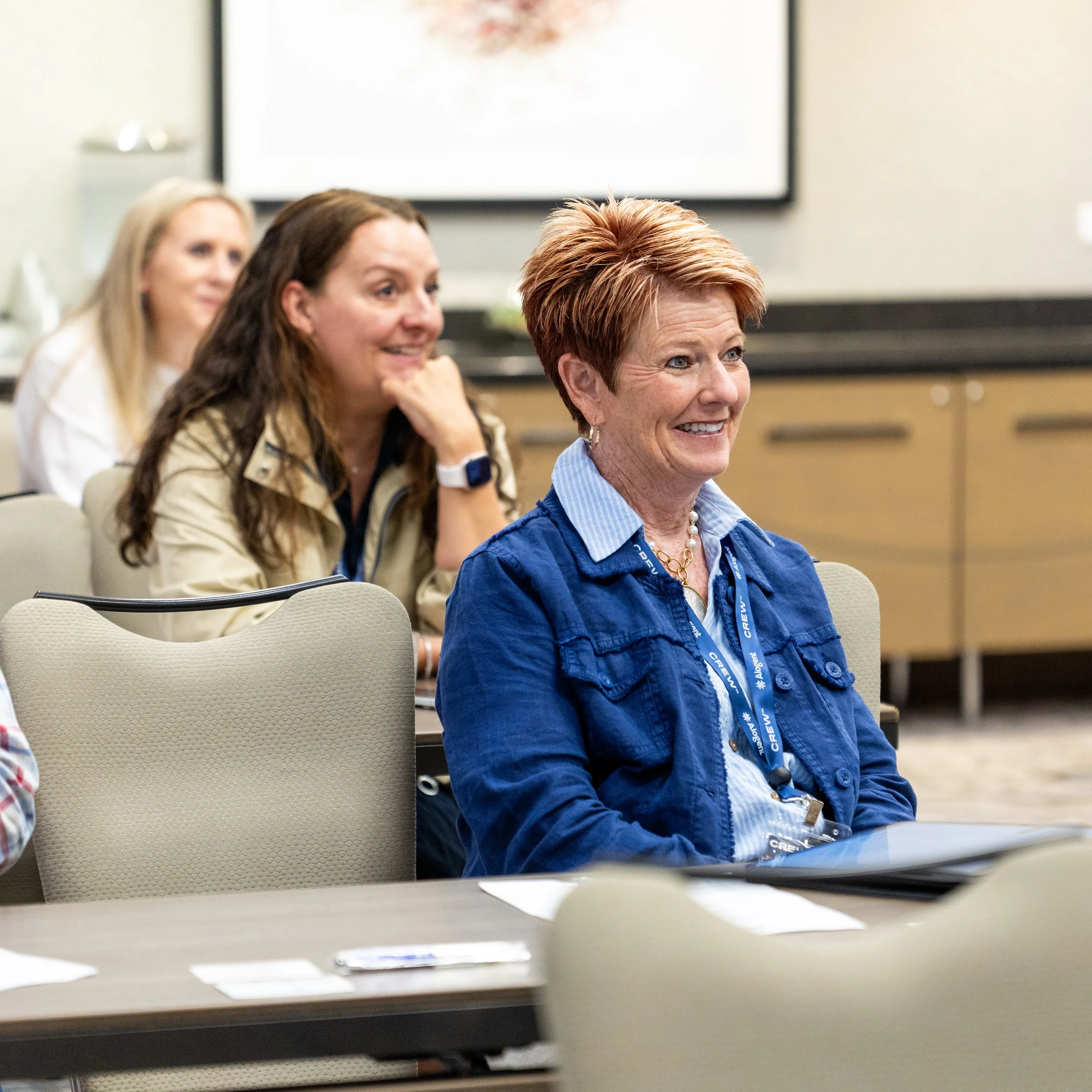 A group of women attending a conference or meeting, seated and smiling, with notebooks and badges around their necks, in a room with a large screen and a table in the foreground.