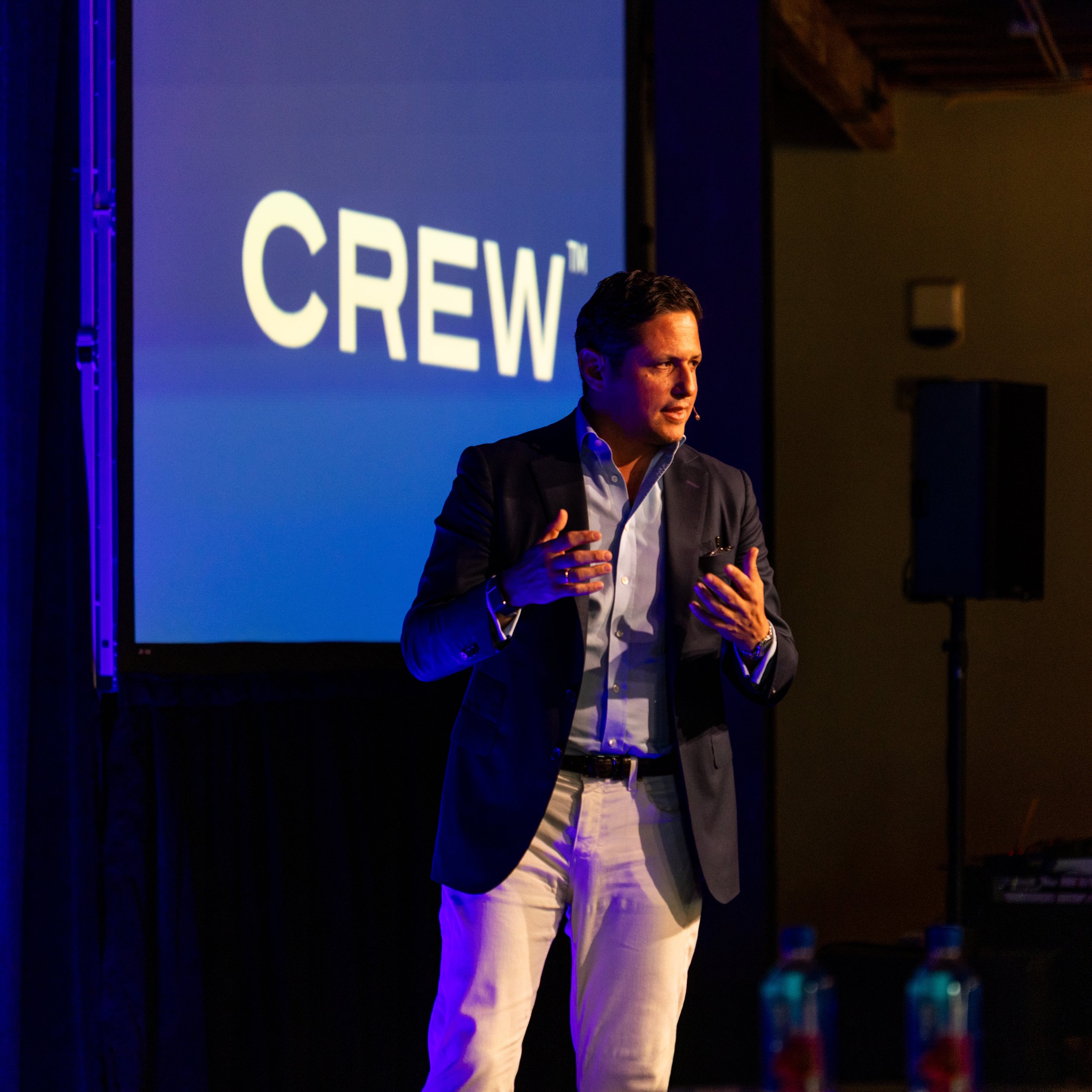 A man in a dark blazer and light-colored pants speaking on stage with a microphone, in front of a blue screen displaying the word 'CREW' in large white letters.