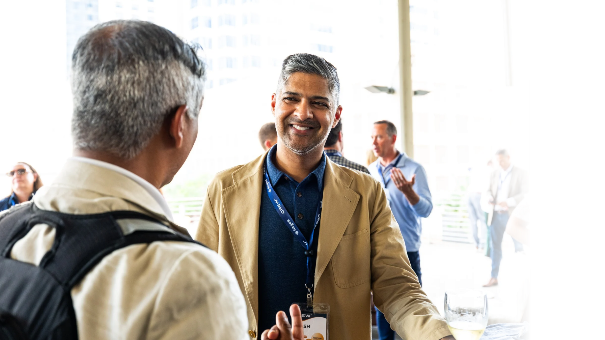 Two men are engaged in conversation at a professional event, with others in the background networking indoors in a bright space with large windows.