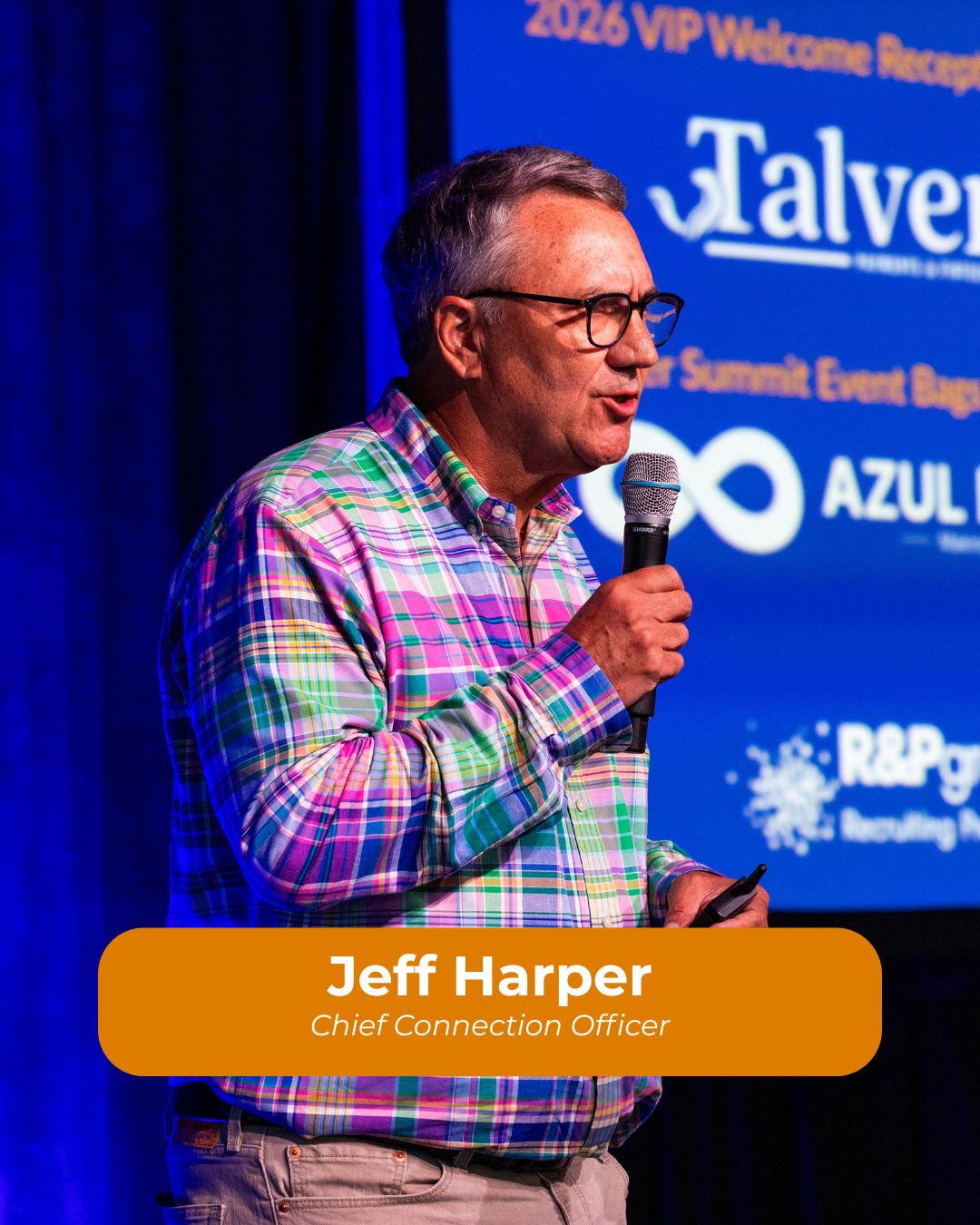 A man wearing glasses and a colorful plaid shirt holding a microphone, standing in front of a blue screen with event and sponsorship information.