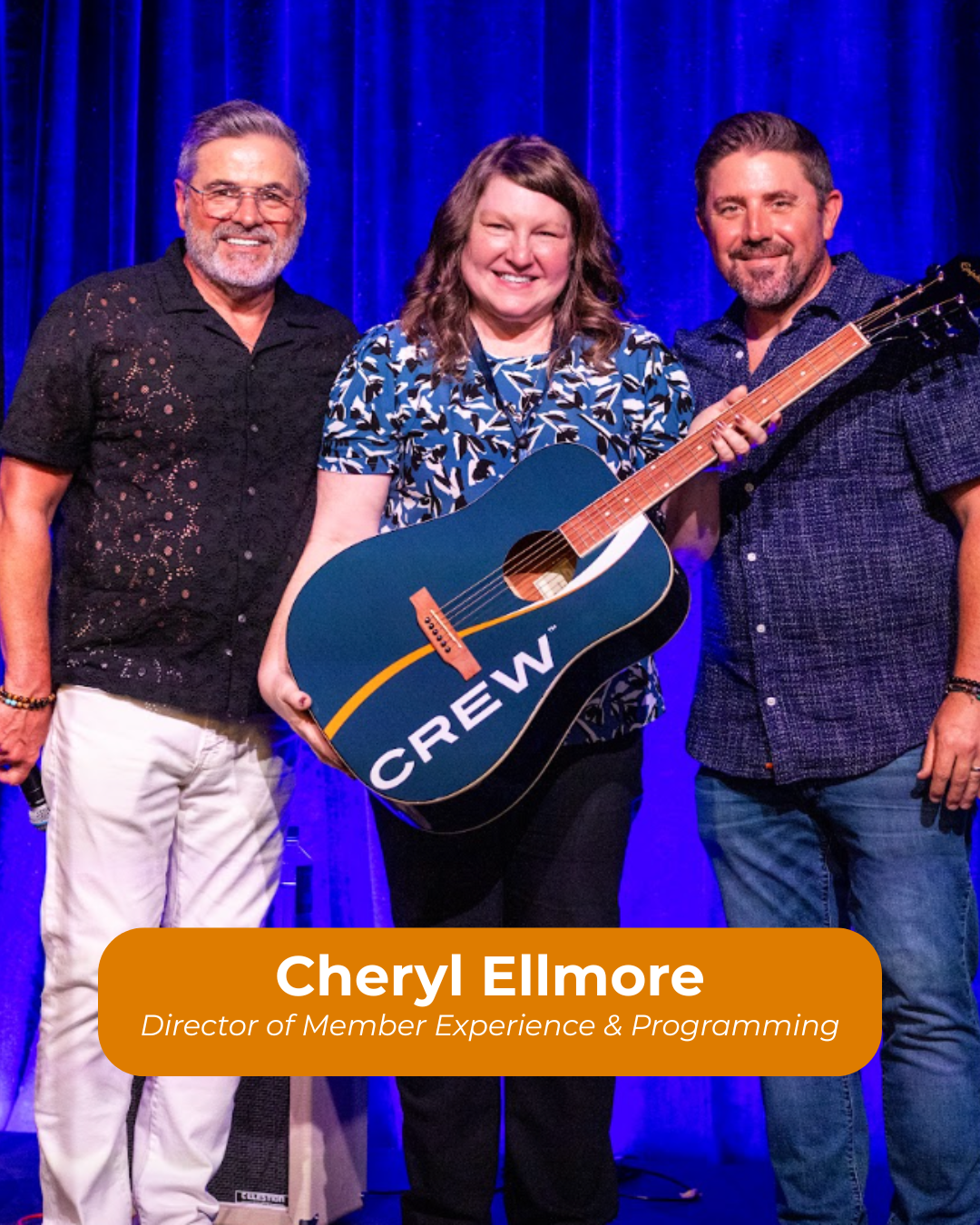 Three people standing on a stage with a blue curtain in the background. A woman in the center holding a guitar with a 'CREW' sign on it. The woman is Cheryl Elmore, identified as the Director of Member Experience & Programming.