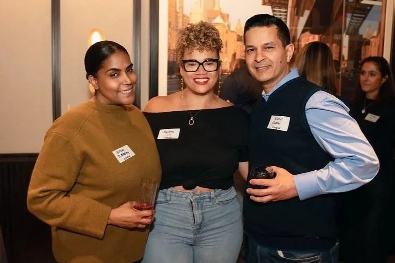 Three people standing close together at an indoor social event, smiling at the camera, with two women and one man, all wearing name tags and holding drinks.