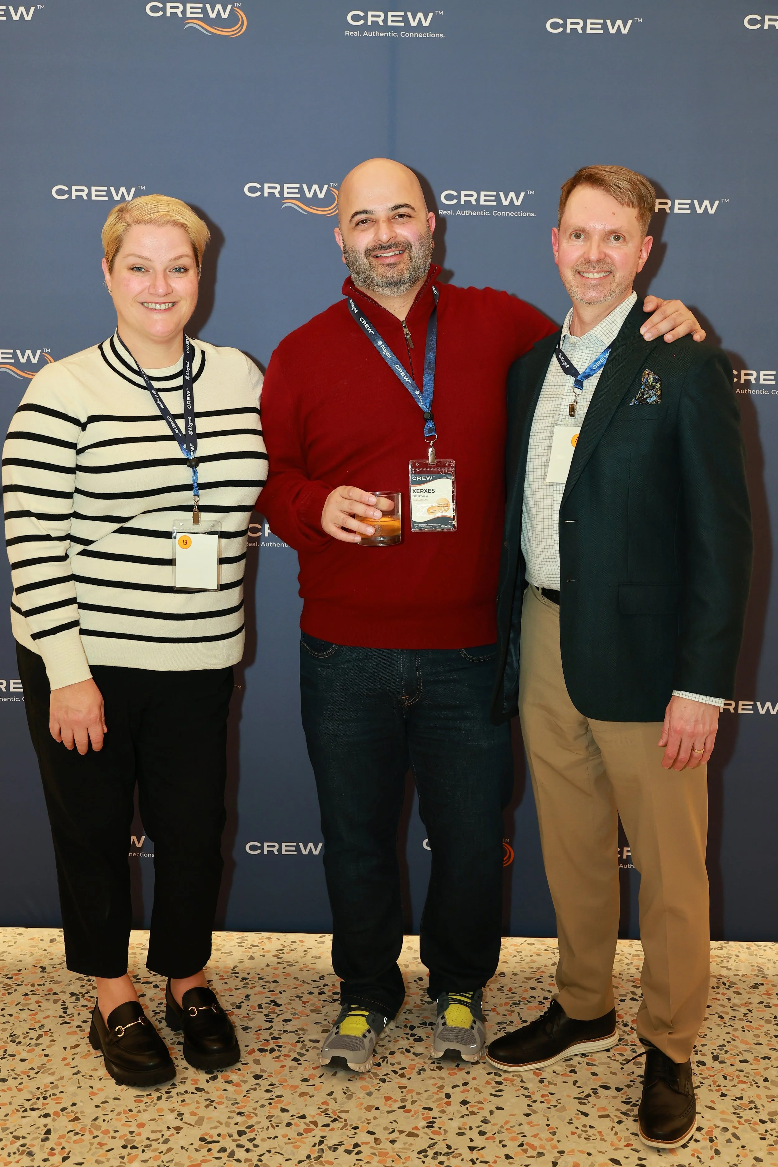 Three people standing in front of a blue backdrop with the word 'CREW' and the tagline 'Real. Authentic. Connections.' They are smiling and wearing name tags. The woman on the left has short blonde hair, is dressed in a striped sweater and black pants. The man in the middle is bald, has a beard, and is wearing a red sweater, jeans, and holding a drink. The man on the right has short light brown hair, is wearing a suit jacket over a checked shirt and beige pants, with his hand on the shoulder of the man in the middle.
