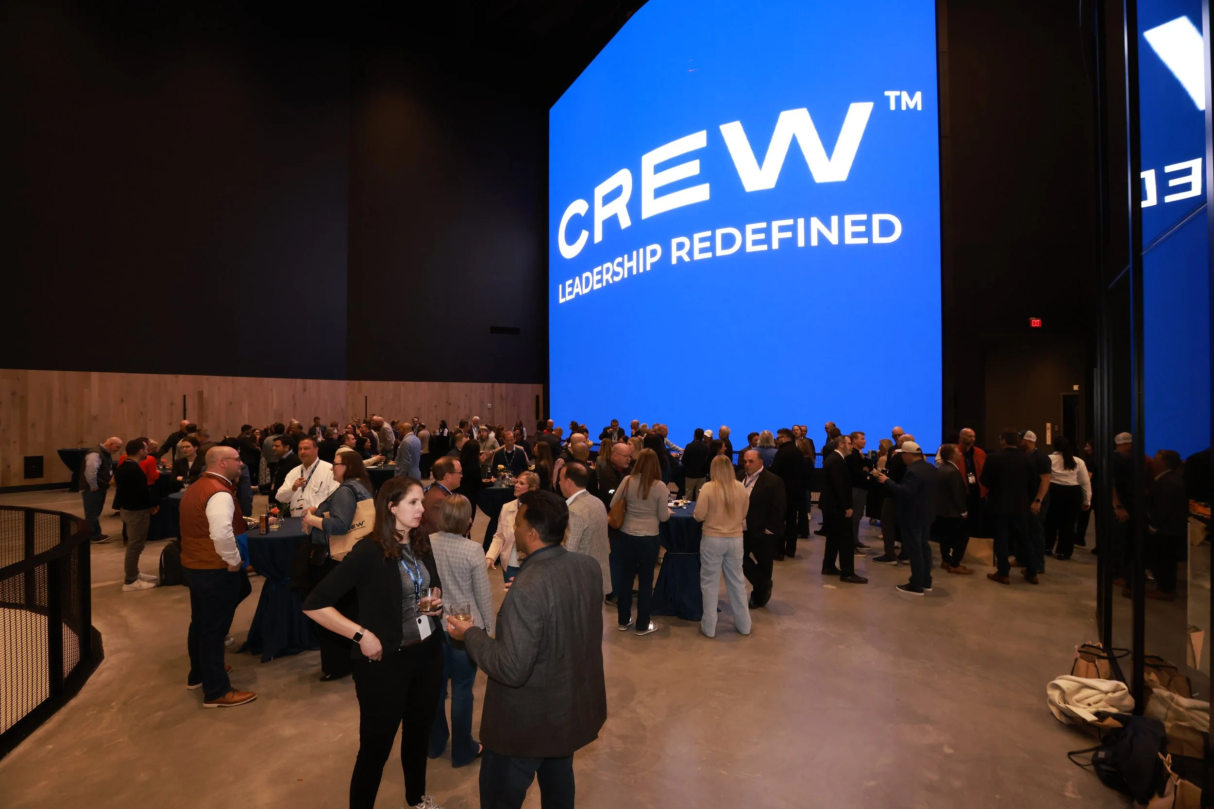 A large conference hall filled with people attending a business event. A prominent blue screen displays the words 'CREW™ LEADERSHIP REDEFINED'.