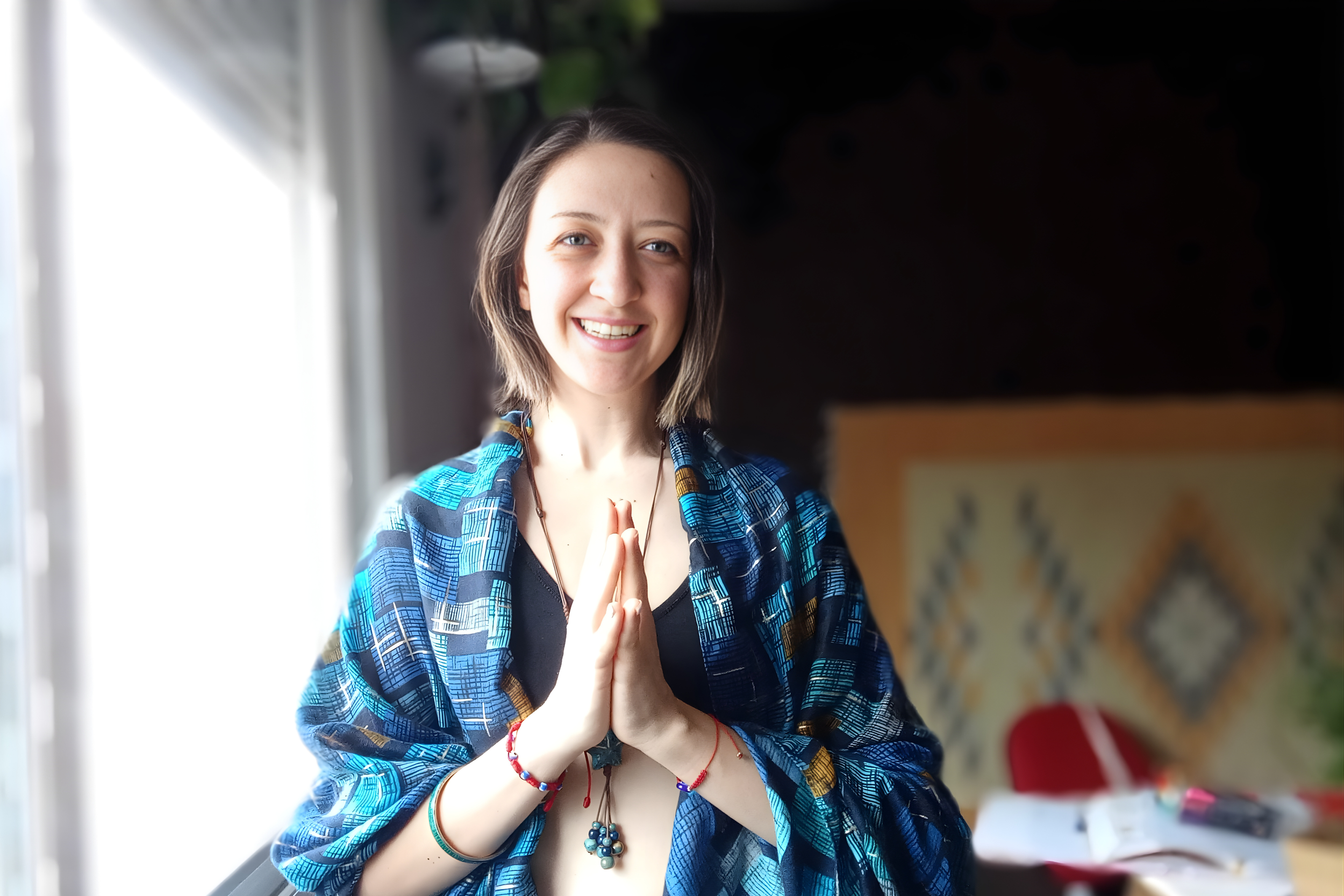 A young woman with shoulder-length brown hair smiling, standing indoors by a window with daylight, wearing a blue patterned jacket and red, green, and blue bracelets, with her hands together in a prayer position.