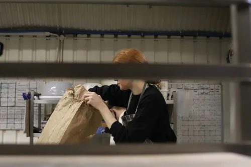 A woman with red hair packing a brown paper bag in a kitchen or industrial setting, viewed through metal bars or a grill.