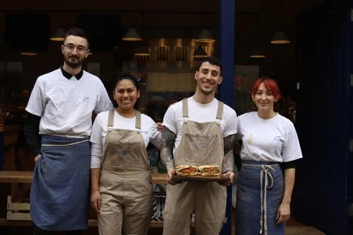 Four restaurant staff members standing outside the restaurant, with one holding a tray of food. They are dressed in chef and staff uniforms, smiling for the photo.