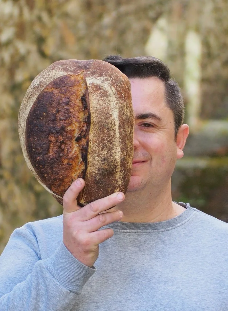 Man holding a large loaf of bread in front of his face outdoors with autumn trees in the background.