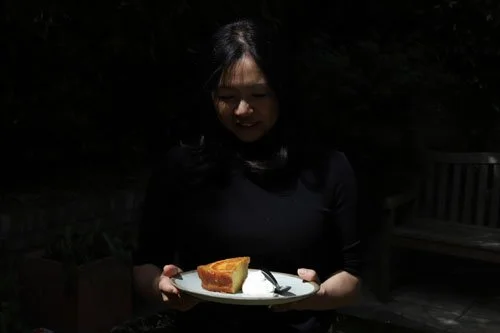 A woman holding a plate with a slice of cake and a dollop of whipped cream outdoors at night.