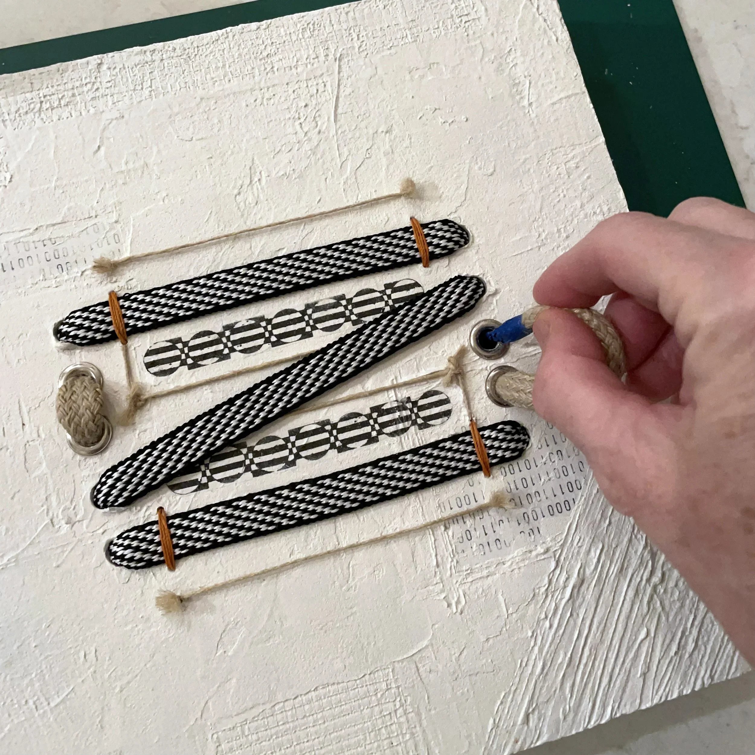 Close-up of a textured white wood panel, with several black and white ropes secured with orange ties, with a person's hand holding vintage twine and grommet.